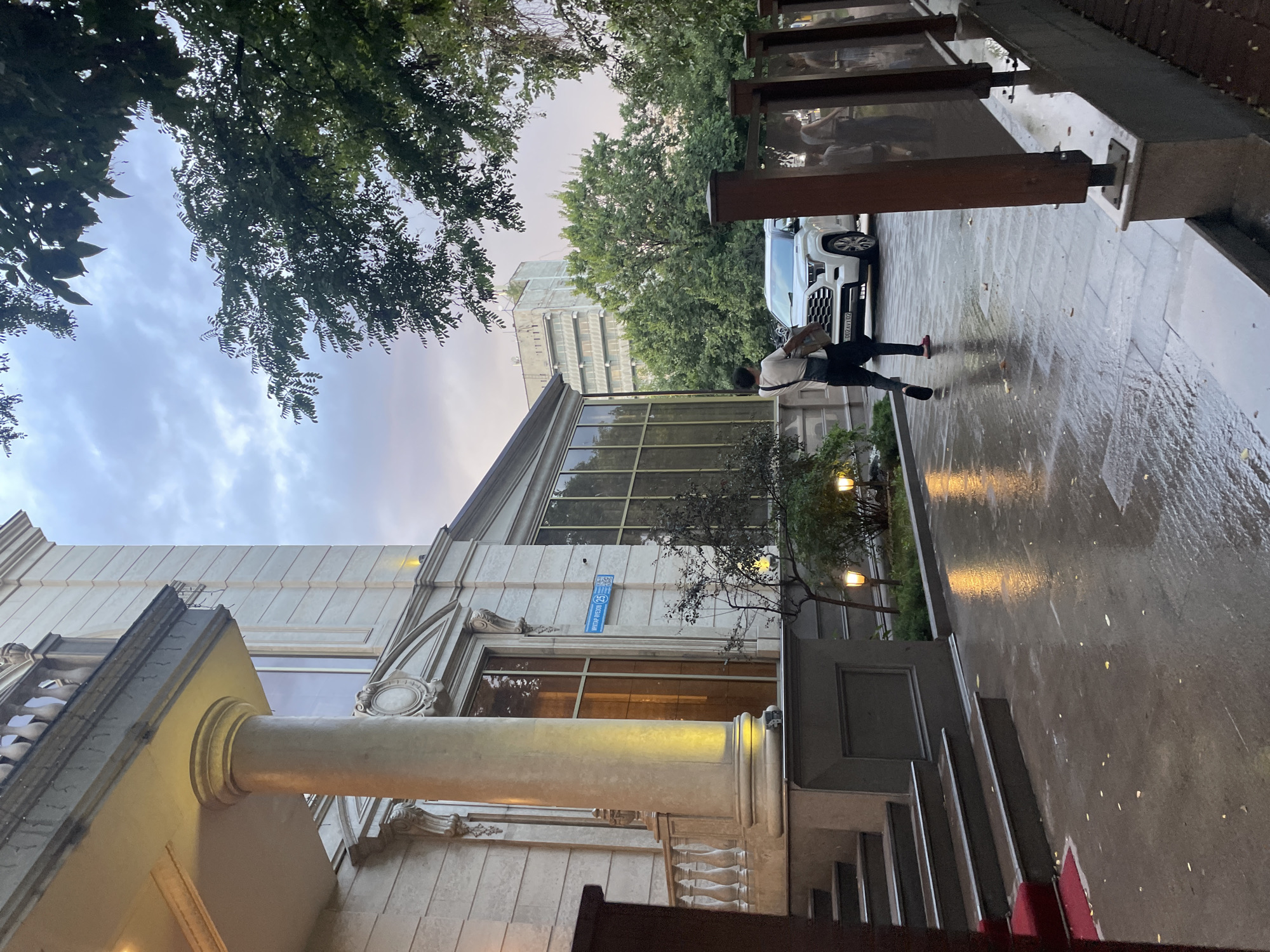 The image depicts the entrance to a building in Almaty, Kazakhstan, on a wet day, likely after rain. In the foreground, a person is walking away from the camera towards a white SUV parked on the paved area. The person is wearing dark pants, a white shirt, and a dark bag slung across their body. The building itself is a large, multi-story structure with classical architectural elements, including large columns and ornate detailing. A red carpet leads up a set of stairs to the entrance. The ground is wet and reflective, with scattered leaves. In the background, trees line the street, and other buildings are visible further in the distance. A street sign with Cyrillic text, "МИКТАР ВУЕЗОН 25", is mounted on the building facade. The sky is overcast with hints of sunset colors.