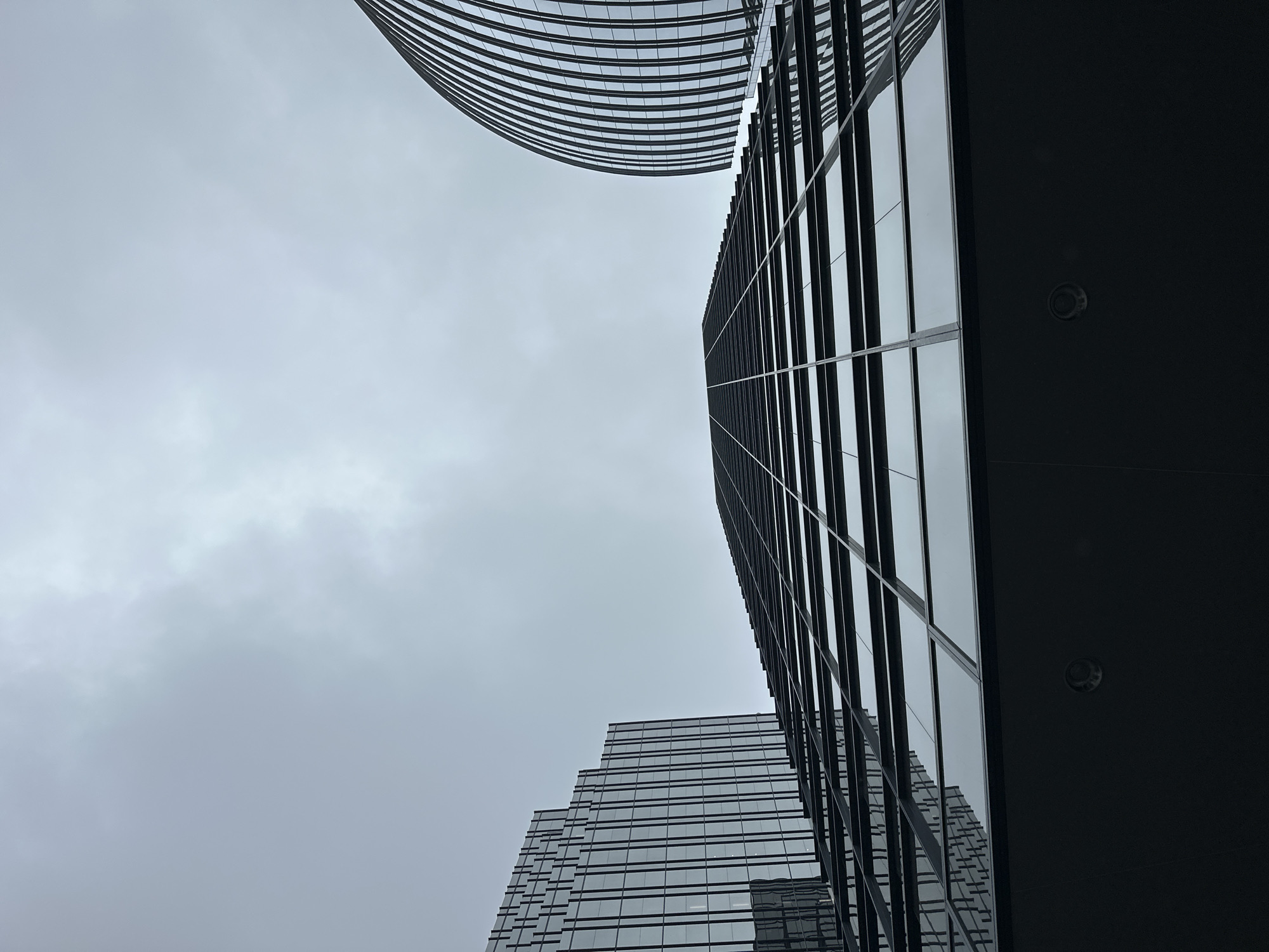 The image shows a low-angle view of three modern skyscrapers against a cloudy sky. The central and largest building features a facade of dark reflective glass, with a grid pattern of horizontal and vertical mullions. To the left, another skyscraper with a similar glass facade has a stepped architectural design. To the right, a third building exhibits a curved, ribbed glass exterior. The foreground is dominated by the dark, solid base of the central building, which has two circular fixtures visible. The overall scene conveys a sense of urban scale and architectural modernity in Burnaby, Canada.