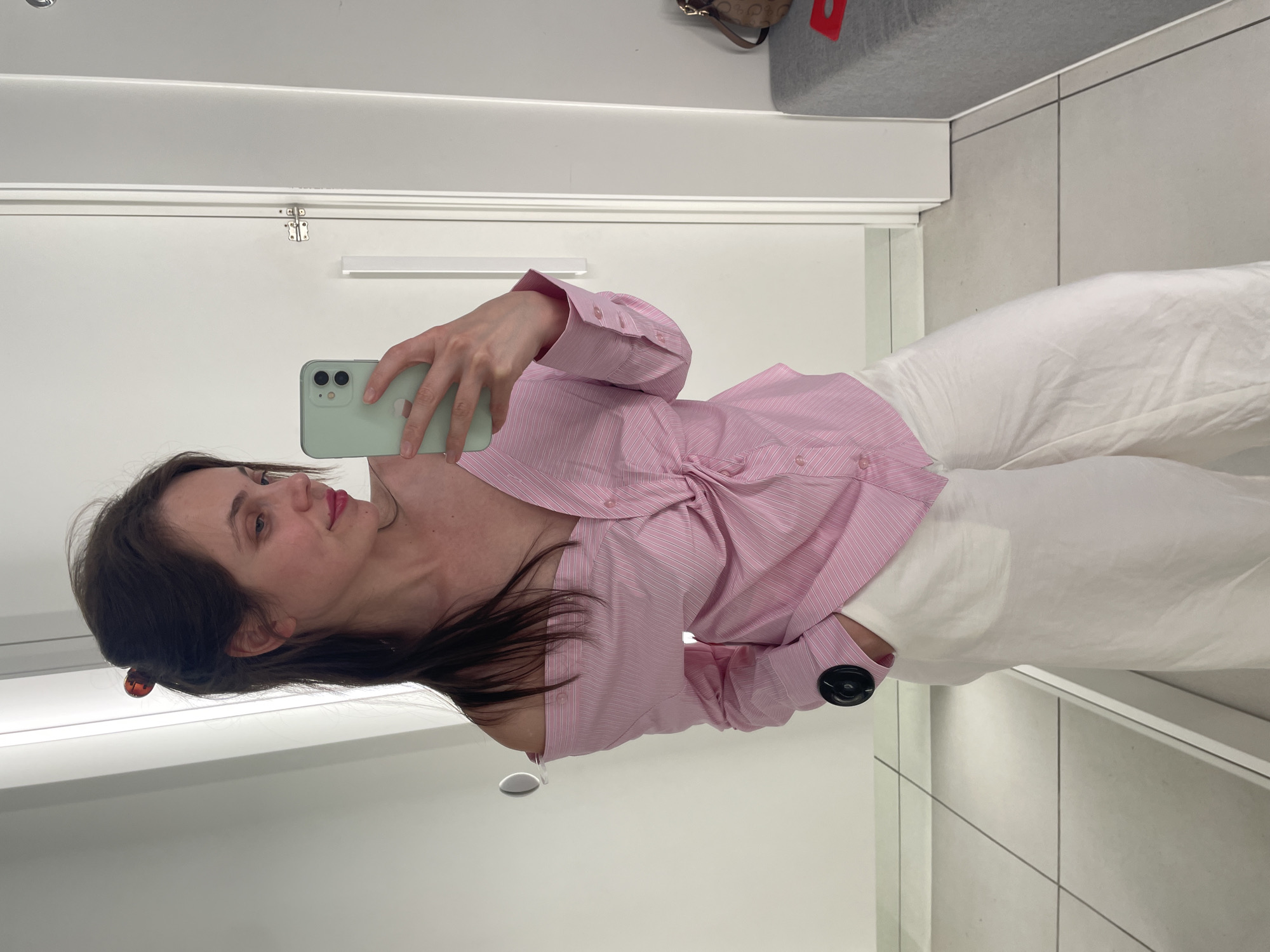 A young woman stands in a fitting room and takes a selfie. She is wearing an off-the-shoulder pink and white striped shirt with a knot detail at the front, and white wide-leg pants. Her hair is pulled back with a brown tortoiseshell clip. She is holding a mint green smartphone in her right hand. The fitting room has white walls and a tiled floor. A grey bench with a brown patterned bag on it is visible in the background. The location is not specified in the image or its context.