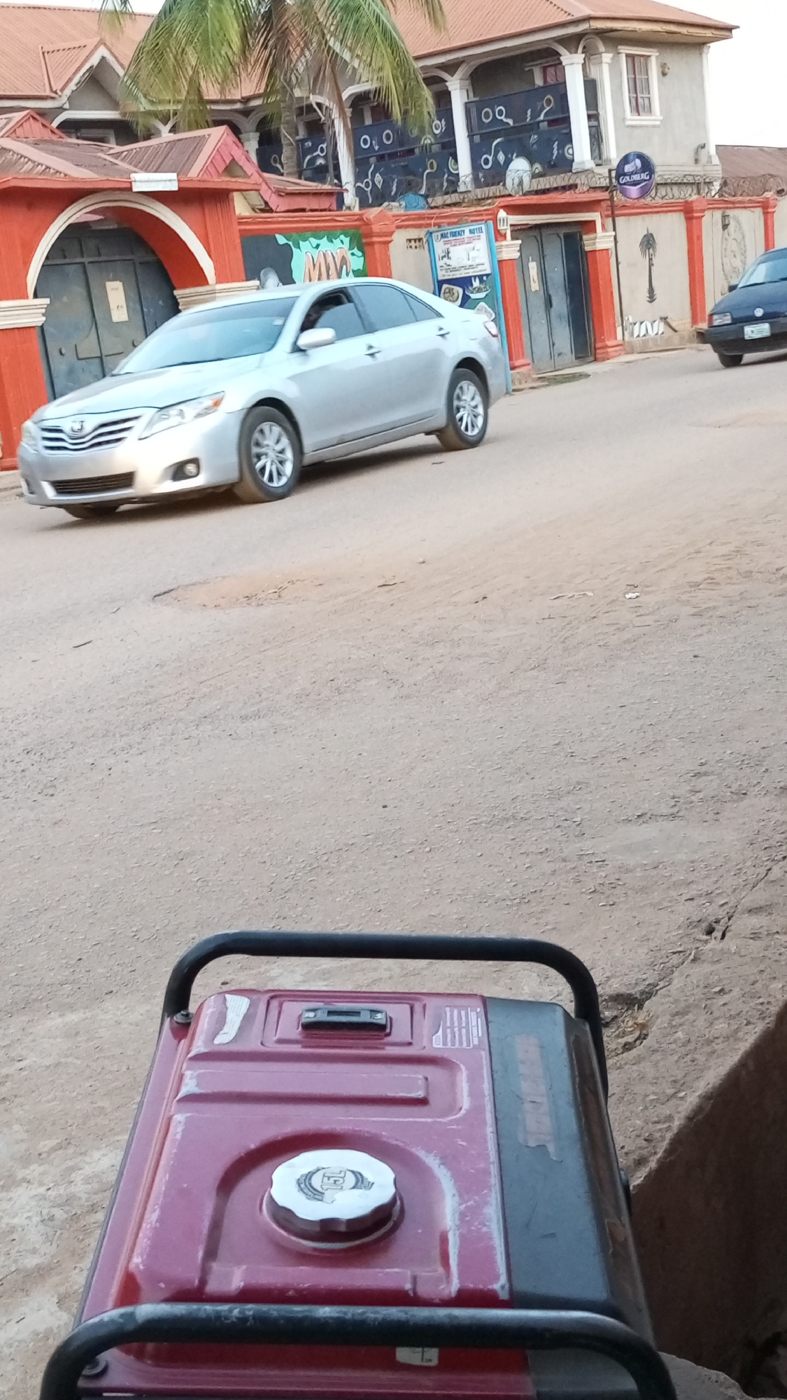 The image depicts a street scene in Sabon Tasha, Nigeria. A silver Toyota Camry is driving on a dirt road. In the foreground, a red generator is visible. The buildings on the side of the road are orange and red with closed doors. There is also a palm tree nearby. Another black car is parked down the street.