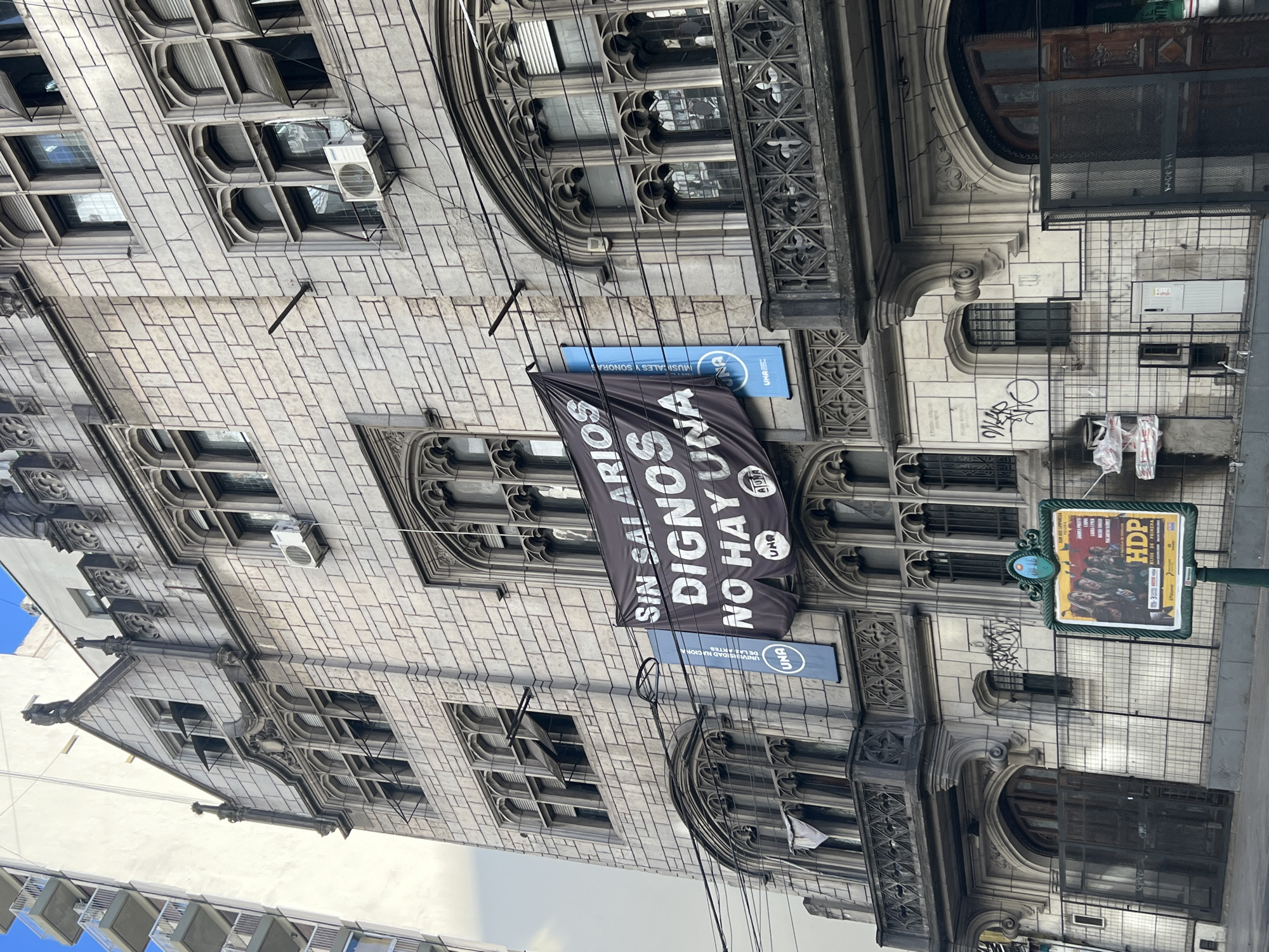 The image shows a stone building in Buenos Aires, Argentina, with gothic-style architectural details. A black banner hangs from the building with the words "SIN SALARIOS DIGNOS NO HAY UNA" ("Without decent salaries there is no one"). A blue sign with the logo "UNA" is visible below the banner. The building has multiple windows with decorative frames and several air conditioning units. A street sign with the abbreviation "HDP" and an image of several people is on the right side of the photo, attached to a green pole. The image is taken from a low angle, showing the building extending upwards.