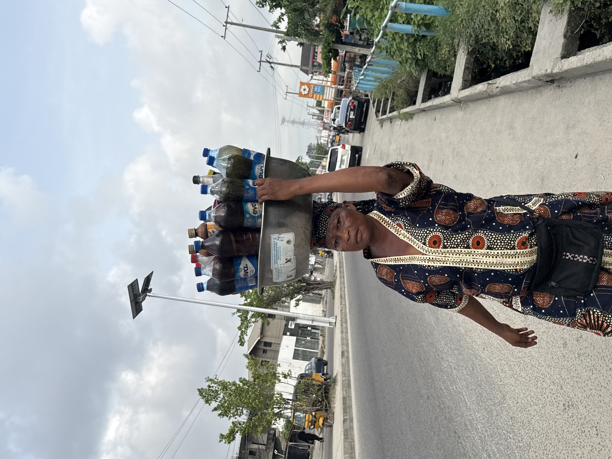 A woman walks on a paved road in Ado, Nigeria, carrying a large metal bowl balanced on her head. The bowl is filled with various bottles of beverages, including water and other drinks. She wears a patterned, dark-colored dress with orange and white accents and a black bag on her hip. The background shows a busy street scene with vehicles, buildings, and trees. Overhead power lines and a solar-powered street light are also visible. The sky is overcast.