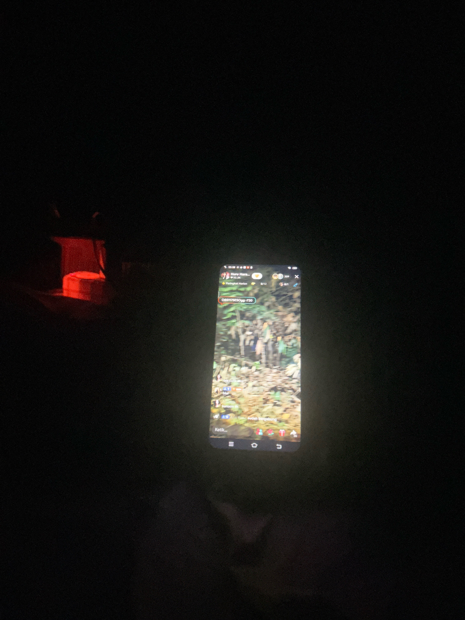 A dimly lit outdoor scene, possibly at night, features a smartphone displaying a live video feed. The video content appears to be a group of people in an outdoor environment with foliage. A red light source is visible in the background to the left of the phone, suggesting an artificial light source or perhaps a distant lamp. The image is dark overall, with the smartphone screen being the primary source of illumination. The location is identified as Cihaurbeuti, Indonesia.