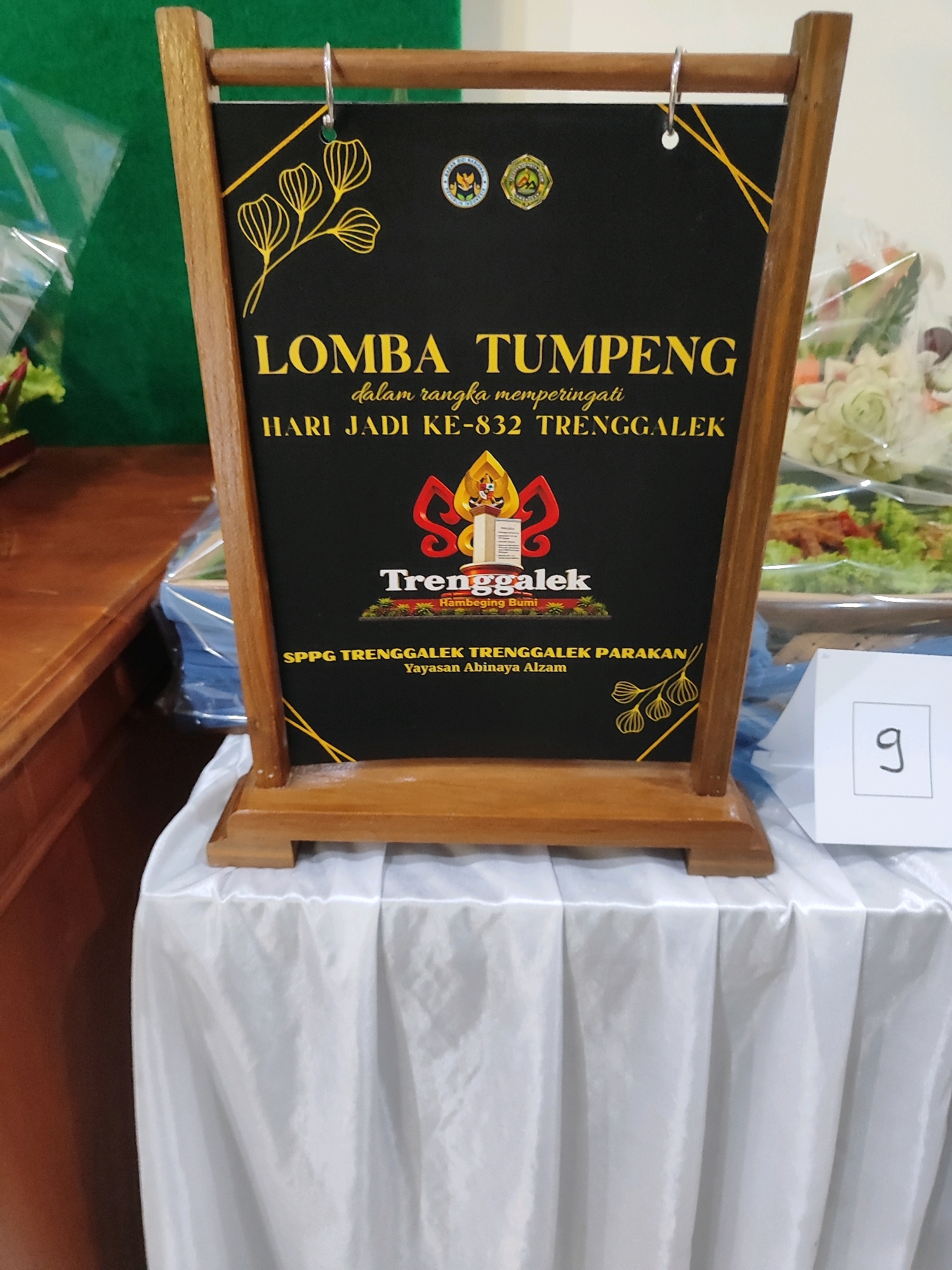 The image shows a black board with text and graphics, mounted on a wooden easel. The text on the board reads "LOMBA TUMPENG dalam rangka memperingati HARI JADI KE-832 TRENGGALEK". Below this, there is a logo with "Trenggalek" written on it, and smaller text below that which reads "Hambeging Bumi". Further down, it says "SPPG TRENGGALEK TRENGGALEK PARAKAN Yayasan Abinaya Alzam". The board is decorated with yellow floral motifs. The easel is placed on a table covered with a white tablecloth. To the right of the easel, there is a small white triangular sign with the number "9" written on it. In the background, on the left, is a green fabric, and on the right, there are food dishes with salads and other items, possibly part of the "Lomba Tumpeng" event. The setting appears to be indoors, possibly a community hall or event space, in Trenggalek, Indonesia, as indicated by the text on the board.
