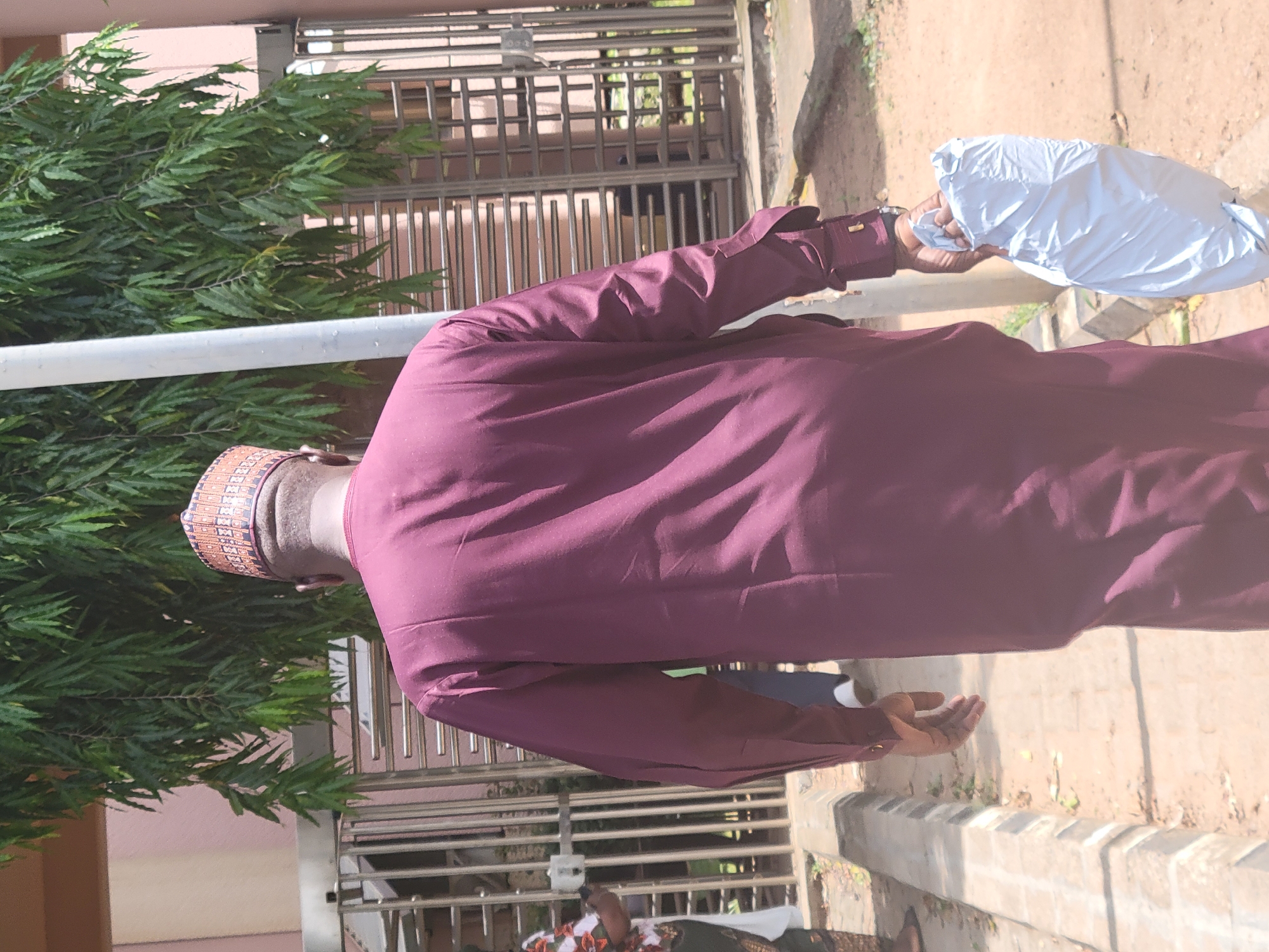 The image, taken in Abuja, Nigeria, shows a man from behind, looking away from the camera. He is dressed in a flowing, long-sleeved maroon garment, possibly a traditional attire. On his head, he wears a patterned cap, likely a kufi, with intricate designs in brown and lighter shades.
In his right hand, the man is holding a white, crinkled package or bag. Another hand, possibly his left, is visible lower down to his side, held open with the palm facing upwards. He appears to be standing or walking outdoors.
The background features lush green foliage, likely from a large bush or tree, to the left and extending across the upper part of the frame. Behind the foliage and to the right, there is a metallic gate or grille structure, suggesting a building or compound entrance. A light pink or beige building facade is also visible behind the greenery. The ground appears to be a light sandy or earthy path, with some sparse vegetation.
The scene is brightly lit by natural daylight, indicating it is daytime. No specific emotions are discernible from the man's posture or the overall scene. There is no visible text on any of the subjects or objects in the image. A small portion of another person, wearing a patterned top, is visible in the lower left background.