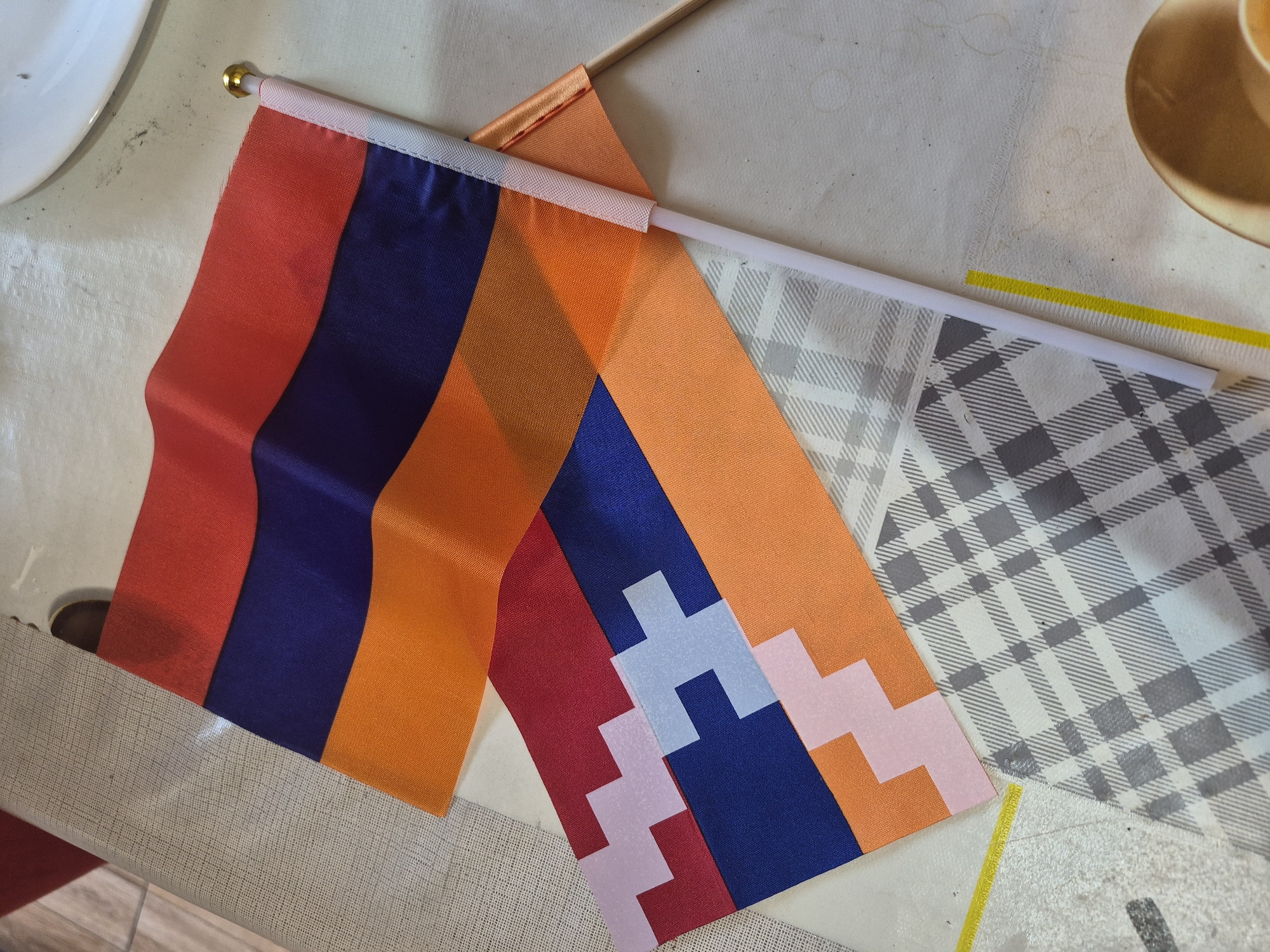 This image shows two small flags. The top flag is the flag of Armenia, with three horizontal stripes of red, blue, and orange. The bottom flag features a geometric pattern in red, blue, and white, resembling a stylized representation of the Artsakh flag. Both flags are attached to white plastic poles, and the Armenian flag has a gold cap on its pole. The flags are laid out on a textured surface, possibly a tablecloth, with a piece of grey and white plaid fabric underneath. Part of a wooden dowel from another object, possibly a third flag or a decorative item, is visible on the right. The image is taken from a top-down perspective. The scene is set indoors on a table, with no identifiable landmarks or context that would indicate a specific location within Yerevan, Armenia.