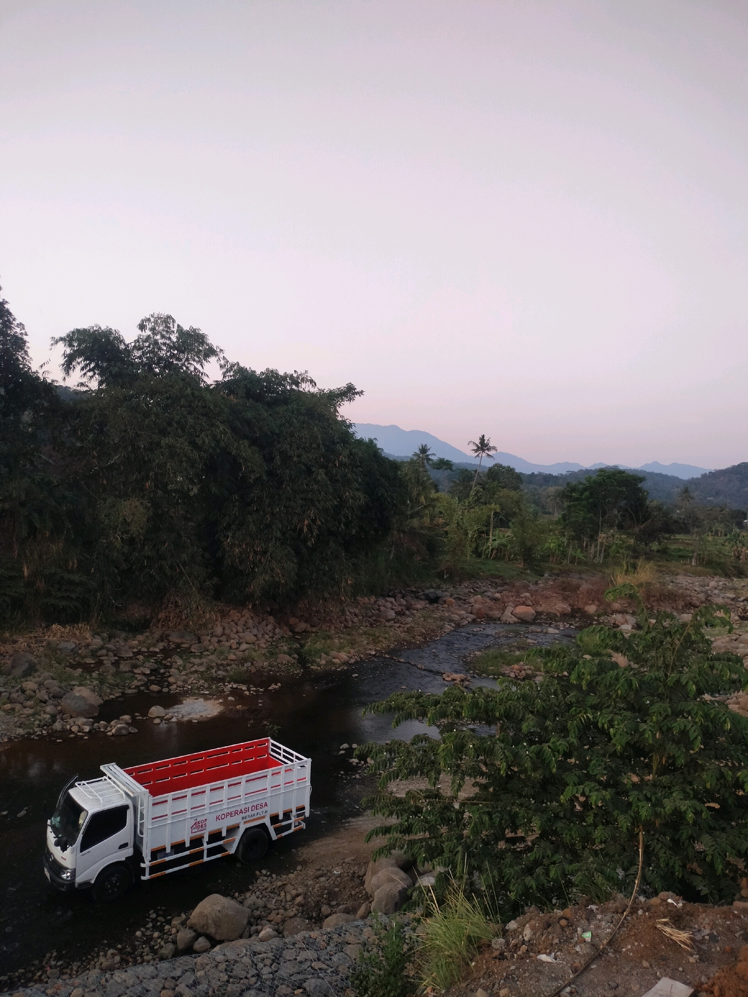 A white dump truck with a red interior is partially submerged in a shallow river. The truck has "KOPERASI DESA MERAH PUTIH" printed on its side. The river flows through a rocky terrain, bordered by lush green trees and vegetation. In the background, rolling hills and mountains are visible under a soft, overcast sky. The scene appears to be in Tersono, Indonesia, suggesting a rural or semi-rural environment.