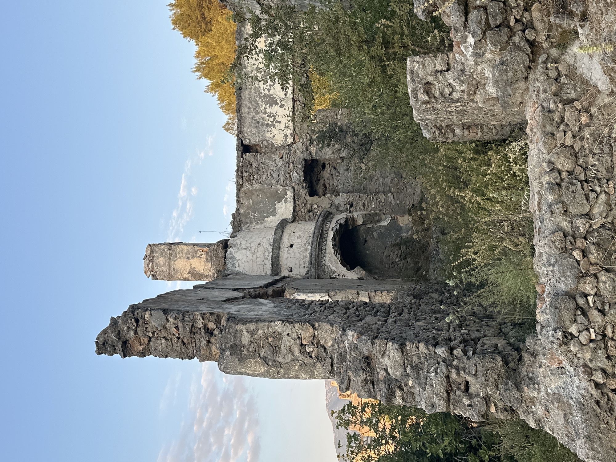 The image shows the ruins of a stone building in Kayaköy, Fethiye, Turkey. The structure is partially collapsed, with weathered stone walls and remnants of plaster. Vegetation, including bushes and tall grass, is growing throughout the ruins and around the exterior. In the background, trees with golden foliage are visible against a clear blue sky with a few wispy clouds. The light suggests it is late afternoon or early evening. There are no people or animals visible in the image.