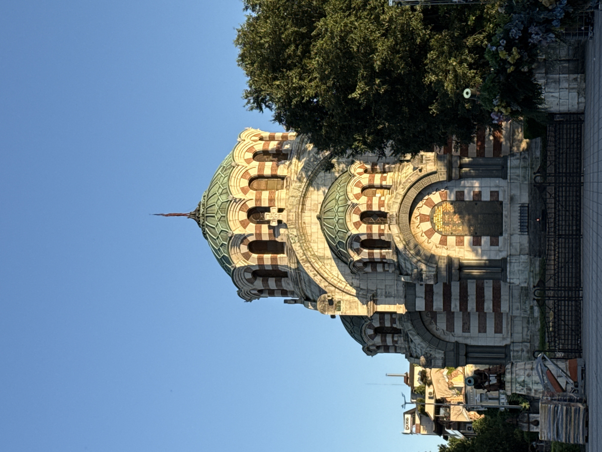 The photograph depicts the St. Nicholas the Wonderworker Russian Orthodox Church, also known as the Russian Chapel, located in Pleven, Bulgaria. The church is a prominent landmark, characterized by its distinctive green domes and striped stone facade, featuring alternating bands of white and red stone. The structure exhibits a classical architectural style with arched windows and decorative detailing.
In the foreground, a black metal fence encloses the church's perimeter. To the left of the fence, a stack of what appears to be construction materials or decorative blocks is visible. Further to the left, a statue of a cannon on a stone pedestal stands as a historical monument. Adjacent to the church on the right, a large, mature tree with dense green foliage dominates the scene, partially obscuring the right side of the building. In the background, several buildings of varying architectural styles and heights are visible, suggesting an urban setting. The sky above is a clear, bright blue, indicative of daytime with fair weather, and the sunlight casts shadows on the church, highlighting its texture and form.
