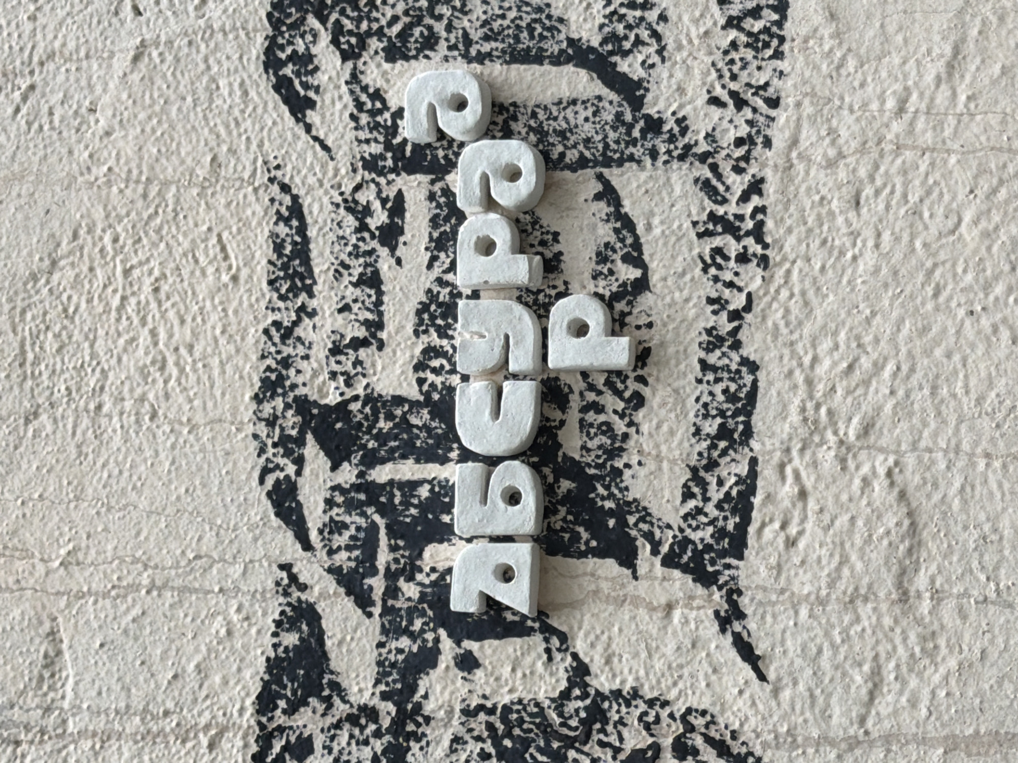 The image shows the word "agenda" spelled vertically with white, rounded letters on a textured surface. The background appears to be a wall with a cream or off-white base and black, abstract markings. The scene appears to be a close-up shot, focusing primarily on the letters and the immediate background. No people are visible. It is not possible to verify the location within the city of Saint Petersburg, Russia from just this image.