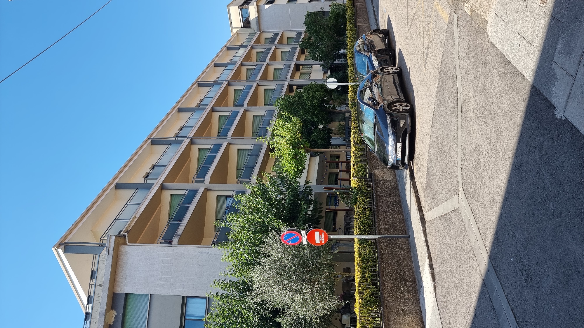 The image depicts a street scene in the city of Olot, Spain, on a clear, sunny day. A multi-story building with balconies runs along the right side of the frame, its facade a light color with darker accents. The balconies have glass railings and are supported by dark columns. Green trees and foliage are visible in front of the building and to the left.

Two dark-colored cars are parked on the street in the foreground, facing away from the camera. The car closest to the viewer has its license plate partially visible, showing "0982 GBL". A traffic sign with two circular prohibition symbols, one red with a blue circle and a red diagonal line, and another red with a white horizontal bar, is positioned on the left side of the street. The street itself is paved with asphalt, with some concrete detailing and pedestrian walkways. The background shows a clear blue sky.