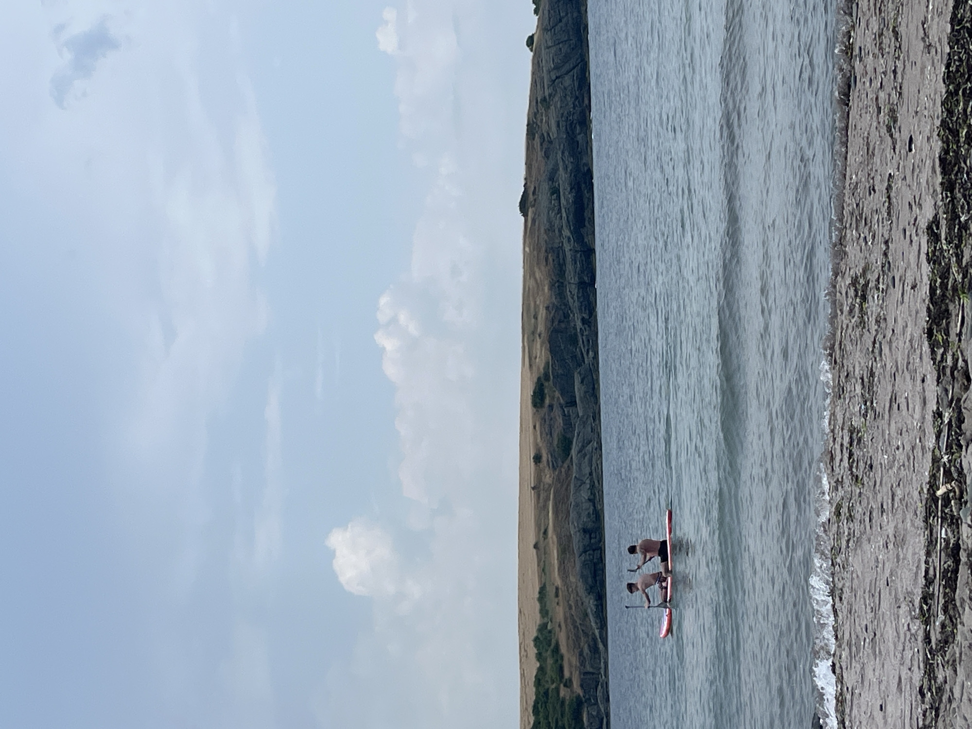 In Ahtopol, Bulgaria, two individuals are paddleboarding on the sea. They are positioned on a single paddleboard, with one person in front and the other behind. Both are actively paddling. The water is calm with small waves. In the background, a rocky coastline with dry, grassy hills rises from the sea. The sky is partly cloudy. In the foreground, there is a beach with sand and scattered debris.