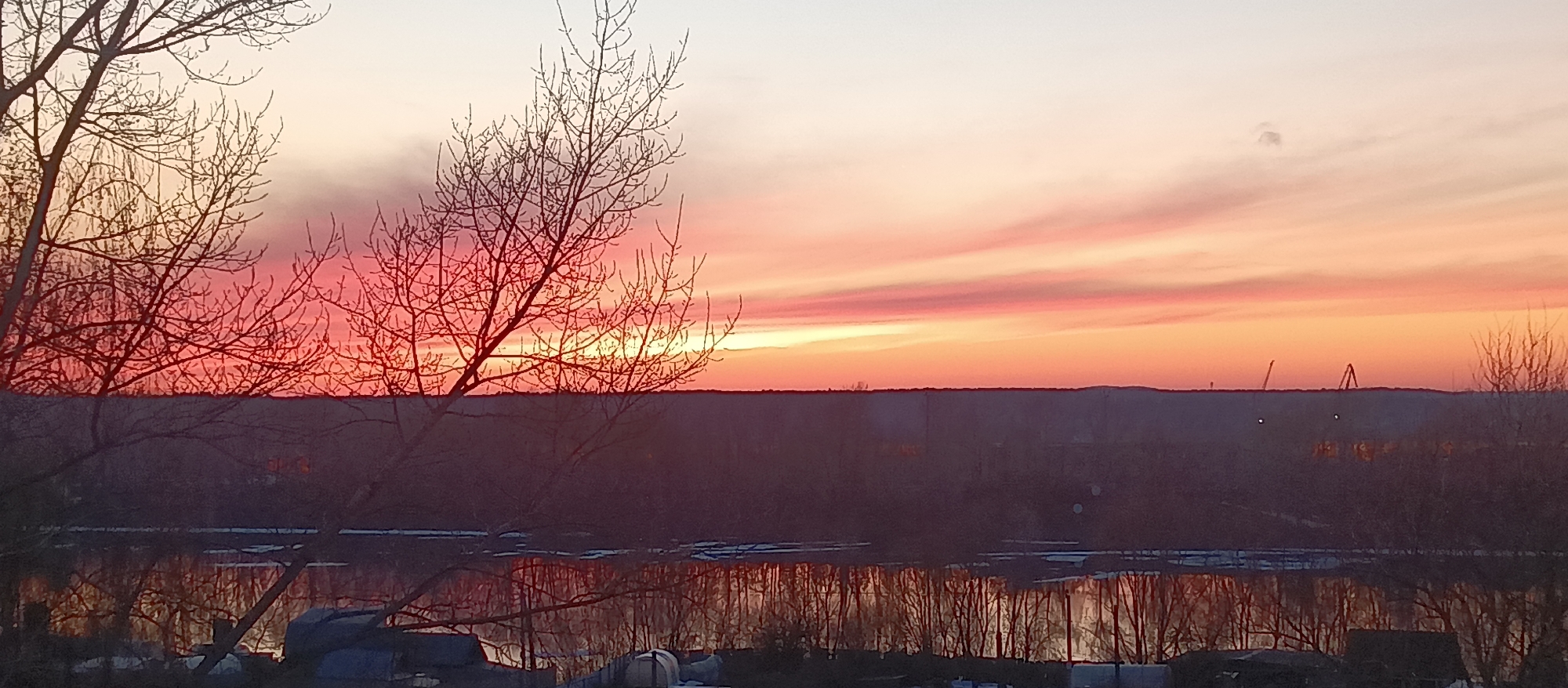The image captures a landscape at sunset, with a prominent river reflecting the vibrant colors of the sky. In the foreground, bare trees stand silhouetted against the sky, their branches intricate and bare. The river reflects the trees and sky, creating a mirror image of the scene above.
Beyond the river, a dark, forested area extends into the distance. On the horizon, there appear to be some structures or industrial elements, including what looks like cranes. The sky is the focal point, with a gradient of colors ranging from pale orange to deep red and pink. The clouds are streaked and layered, adding depth and texture to the scene.
The lighting indicates either dawn or dusk, with a warm, soft glow illuminating the sky and reflecting in the water. The overall mood of the image is serene and peaceful. The exact location within the city of Unknown, Unknown is unclear without further information.