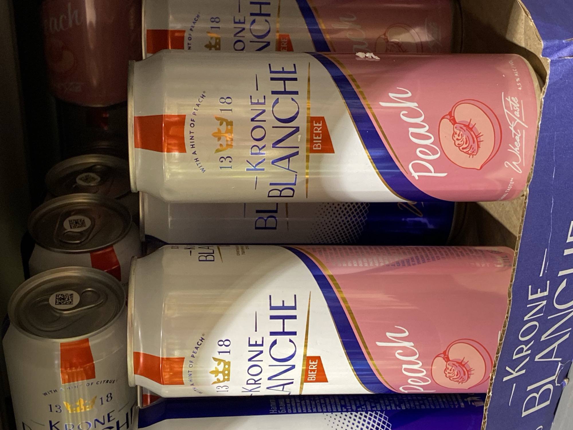 The image displays an assortment of alcoholic beverage cans, primarily of the "KRONE BLANCHE BIERE" brand, stored within a brown cardboard box. The cans are packed closely together, some upright and others on their sides, suggesting a storage or display context in Votkinsk, Russia.
Two main flavor varieties are prominent:
1. **Peach**: These cans feature a pink background with a white "Peach" script and an illustration of a peach. Text visible on these cans includes "4.5% ALC/VOL", "A Wheat Taste", and "WITH A HINT OF PEACH\*".
2. **Citrus (implied)**: Other cans are predominantly white with orange accent bands. Partial text on these indicates "WITH A HINT OF CITRUS".
All cans prominently display "KRONE BLANCHE BIERE" in blue and gold lettering, along with the numbers "13/18" or "13 18". The word "BIERE" (French for beer) is clearly visible.
Various Cyrillic inscriptions are present on the cans, reinforcing the Russian location context. These include "Напито Бланш" (Drink Blanche), "СОСТАВ: Вод" (Ingredients: Water), "КАРАМЕЛЬНО" (caramel), "АЛКОГОЛЬ" (alcohol), "БРОЖЕНИЯ" (fermentation), "ЭКСТРАКТ-" (extract), "ОРИГИНАЛЕ" (original), and "ЗАВОД" (factory).
No people are visible, and no specific event or activity is taking place beyond the storage of these products. The lighting appears to be artificial, typical of an indoor setting like a warehouse or store.