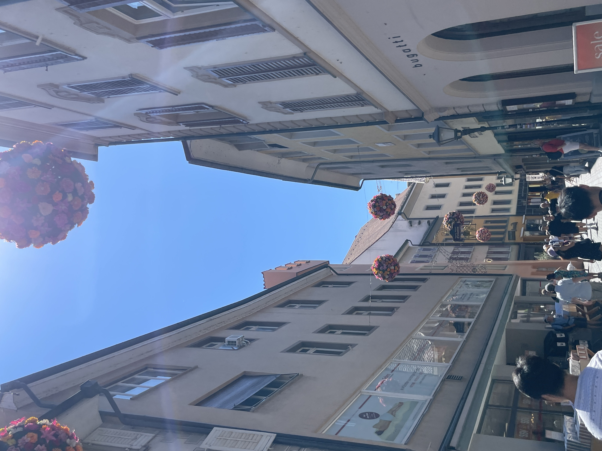 In Constance, Germany, on a bright sunny day, a street scene unfolds, featuring buildings with traditional European architecture. Flower-laden spheres are suspended across the narrow street, serving as decorative elements. On the left side of the street, a building showcases several storefronts with large windows displaying merchandise and advertisements. An air conditioning unit is visible on the upper facade of this building. On the right, another building features arched windows and the prominent "bugatti" logo, suggesting a retail establishment. A lamppost stands in front of this building. Several people are walking along the street, some appearing to be tourists or shoppers, engaged in casual activities. The sky is clear and blue, indicating good weather.