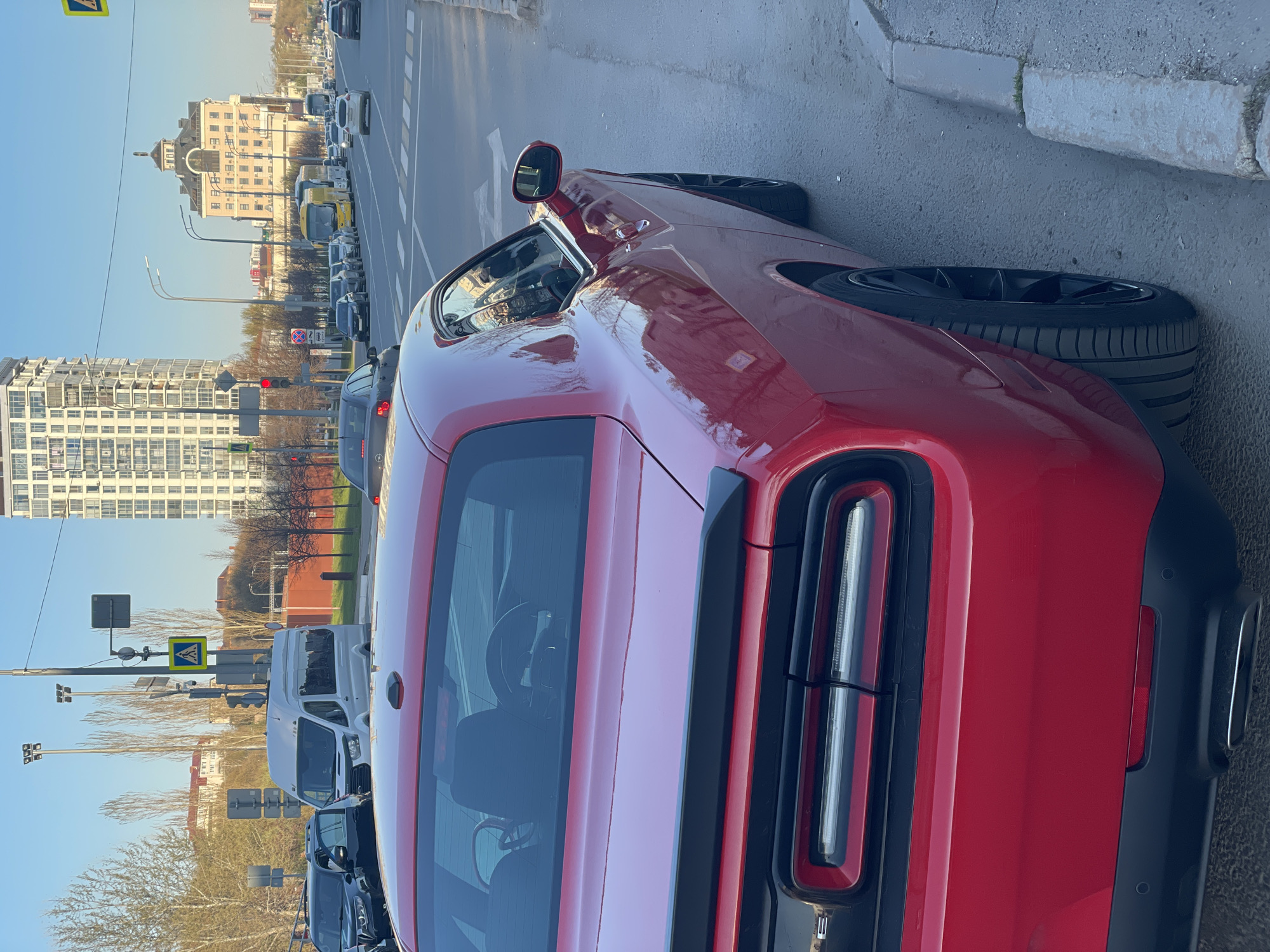A close-up, angled shot depicts the rear of a red Dodge Challenger parked on the side of a street. The car's taillights, trunk, and rear bumper are visible. The background reveals a cityscape, including buildings of varying heights and architectural styles, typical of Kazan, Russia. A street is filled with cars. Street signs and bare trees line the side of the road, and the sky is pale blue.