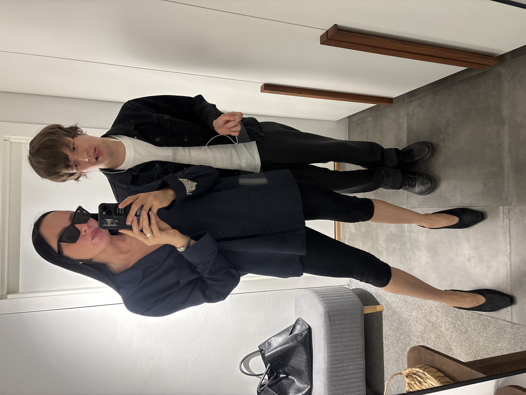 The image shows two people in what appears to be a changing room, possibly in a store. A woman with dark hair and sunglasses is taking a selfie with her phone. She's wearing a dark jacket and black leggings, with black ballet flats on her feet. A gray padded bench is visible behind her, with a black handbag resting on it. A young man stands behind her, wearing a white T-shirt, black jacket, and black pants, with black shoes. He has an earbud in his ear. The room has white walls and a gray floor, with wooden fixtures. The city of Chaikovsky, Russia is not directly visible. There is no information to conclude the image was taken there.