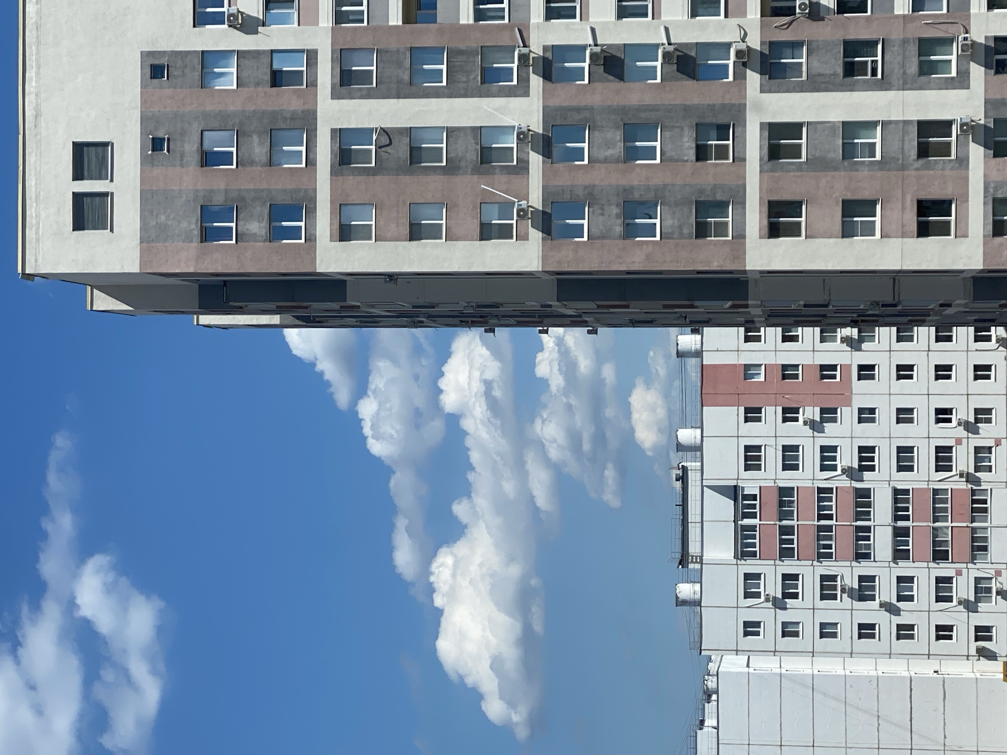 The image depicts a contemporary urban landscape with apartment buildings under a clear blue sky dotted with fluffy white clouds. The buildings are modern, featuring geometric designs and varying color palettes. One prominent building on the right displays a vertical stripe pattern in shades of grey and beige, punctuated by numerous windows and air conditioning units. To the left, a slightly shorter apartment building is visible, characterized by a lighter facade with pink and white accents, also fitted with air conditioning units. The setting appears to be a residential area in Ulyanovsk, Russia, suggesting a typical city view.