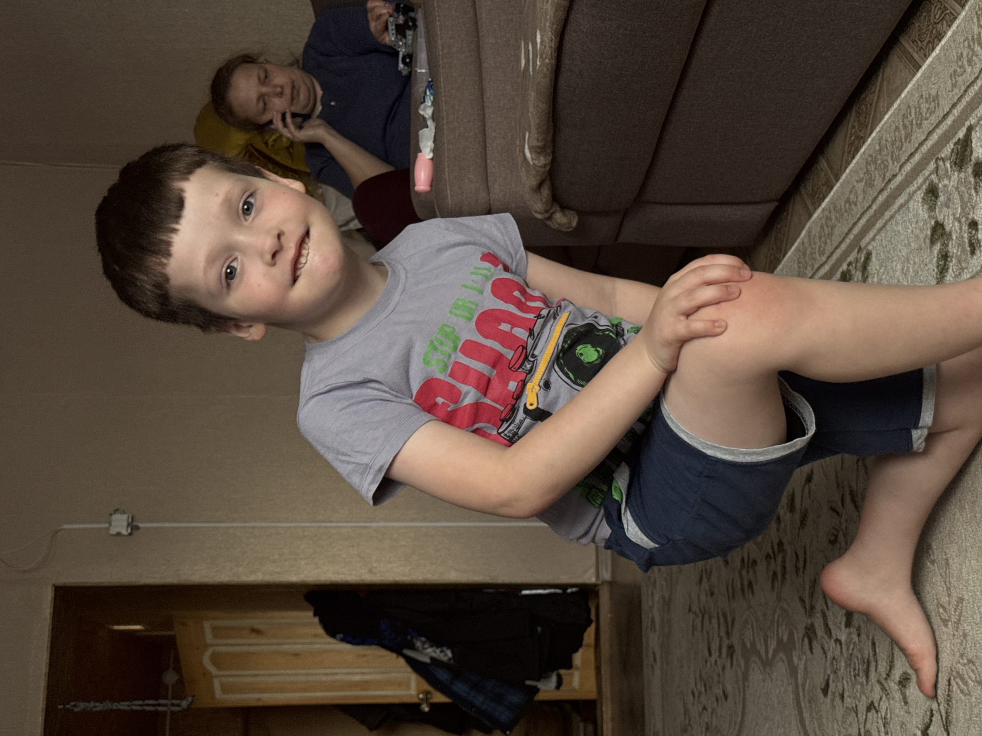 A child is lying on the floor, smiling and holding his knee. He is wearing a gray t-shirt with a red graphic and blue shorts with gray trim. An adult is visible in the background, sitting on a brown couch and talking on a cell phone. A patterned rug is on the floor, and a cabinet is visible in the lower left corner. The location is Кемуль, Russia.