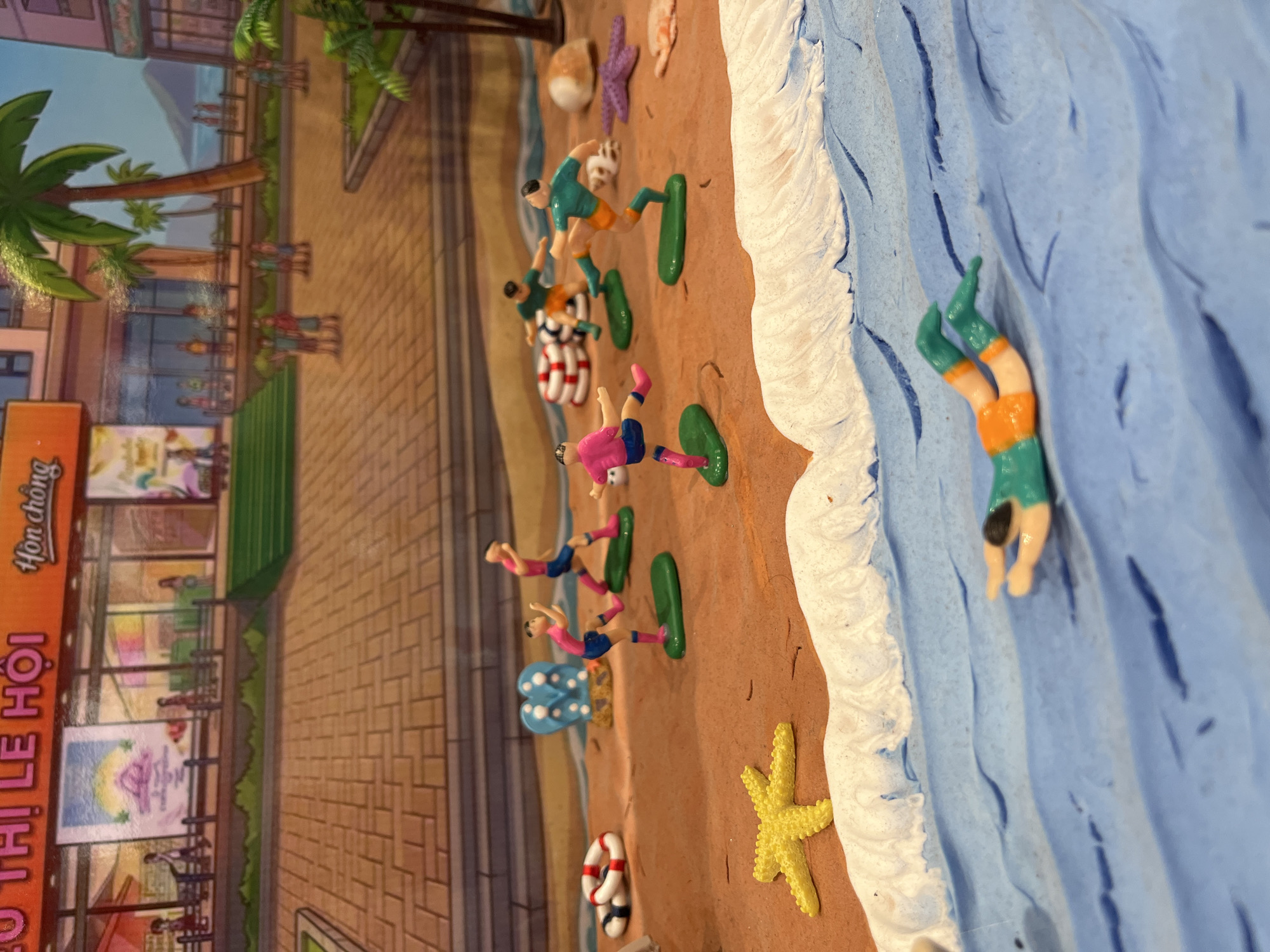 A miniature diorama depicts a scene at a beach in Bac Nha Trang Ward, Vietnam. Clay figures are positioned on a sandy beach area with an adjacent blue clay ocean. Some figures appear to be sunbathing on green platforms. One figure is diving into the ocean. A building facade with signage is located on one side. Details like palm trees, starfish, shells, and beach-themed objects such as a life preserver are included. The level of detail and perspective suggest a staged or constructed scene.