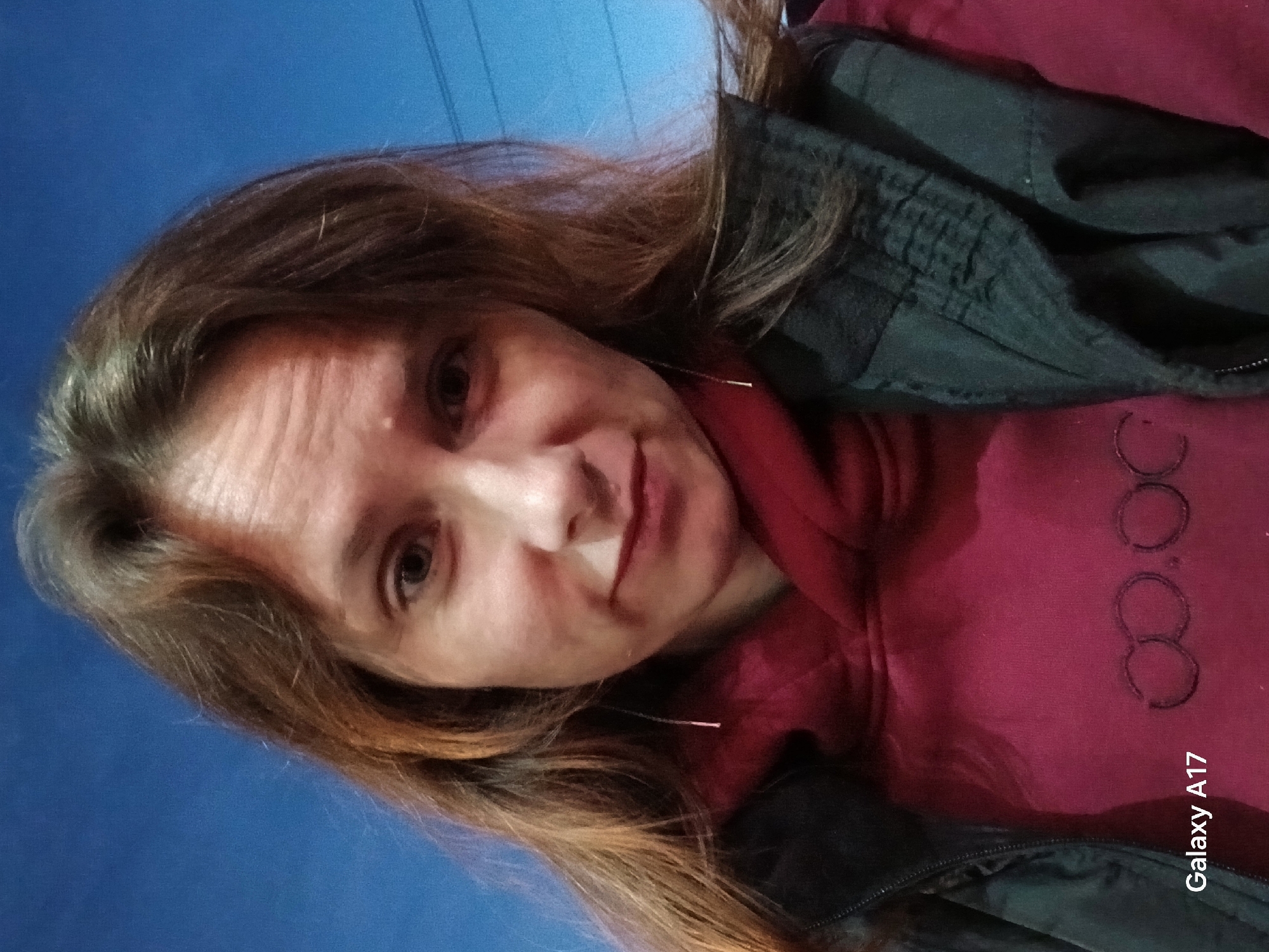 Here is a description of the image:
A woman is taking a selfie. She is wearing a burgundy hooded sweatshirt and a black jacket. The background is blue, and there appear to be wires in the upper portion of the frame. "Galaxy A17" is visible in the lower right corner, which is likely a watermark from the phone used to take the picture. The exact location in Yekaterinburg, Russia, cannot be determined from the image alone.