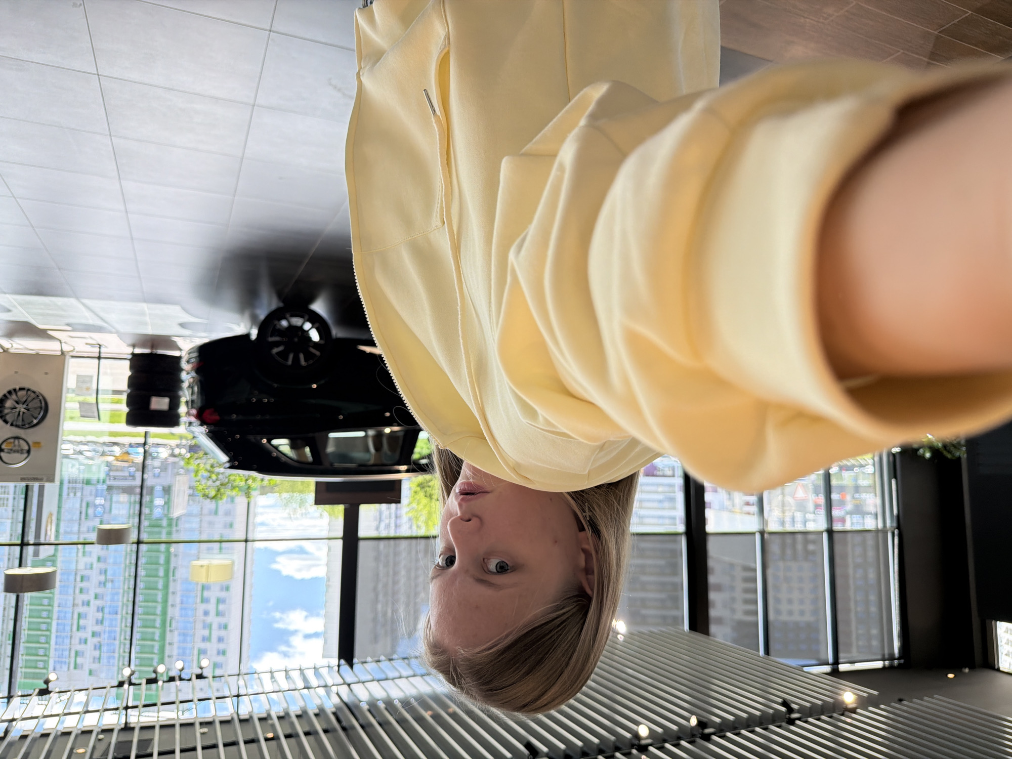 A woman is shown in a selfie taken indoors, upside down. She is wearing a light yellow jacket. Behind her is a modern interior with large windows reflecting the cityscape. A car and tires are also visible in the reflection. Based on the window reflection, the location appears to be in Izhevsk, Russia, based on the architecture and urban layout. The primary subject is the woman in the foreground; the background suggests a showroom or display area.