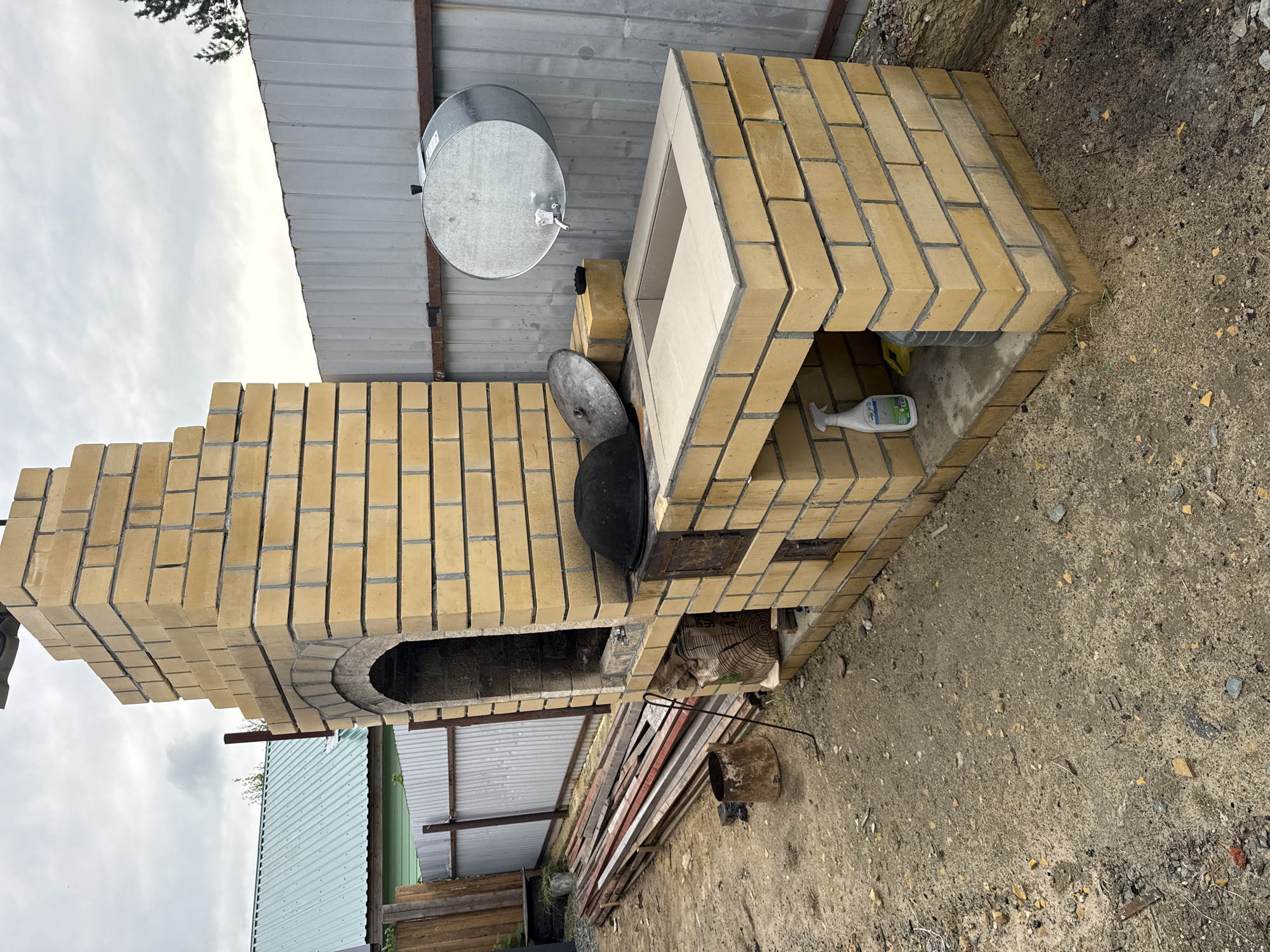 This is a medium-shot, eye-level, outdoor photograph of a newly constructed brick barbecue grill and oven in Sovetskiy, Russia. The barbecue is made of yellow brick and features a tall chimney with an arched opening. To the right of the chimney is a flat surface with a square opening, possibly for a stovetop or sink. Below this surface, there is an open space with a bottle of cleaning spray and a yellow container. A large, round metal water tank with a spigot is mounted on a metal fence behind the barbecue. In front of the barbecue, a pile of wooden planks and a rusty bucket are visible. The ground is covered in gravel and dirt, and a grey corrugated metal fence borders the property. The sky is overcast. The overall scene suggests an unfinished outdoor kitchen area.