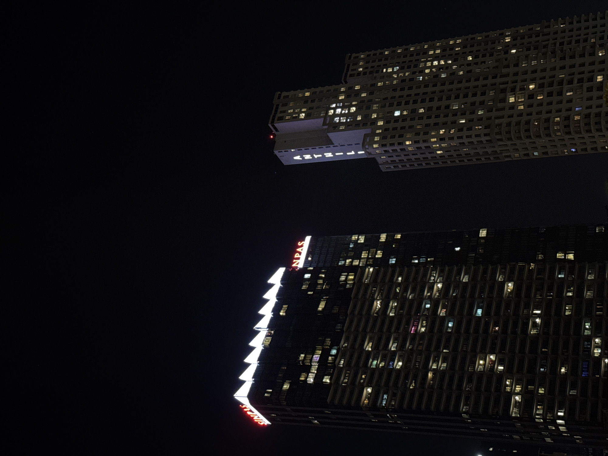 Two modern buildings are silhouetted against a dark night sky. The building in the foreground is a tall, dark skyscraper with many windows illuminated from within, showing hints of various colored lights. The building has a distinctive jagged, white illuminated edge running down its side, and red neon signage spelling out "SINPAS" at the bottom and top. The building in the background, which appears to be farther away and possibly higher, is lighter in color and also features many illuminated windows. It has a distinctive, stepped architectural design and text that appears to read "ANTELIA" in white illuminated letters on its lower facade. The scene suggests an urban setting at night, possibly in Şişli, Turkey, given the location context. There are no people visible, but the lit windows imply occupancy and activity within the buildings. The overall mood is one of nighttime urban vibrancy and architectural presence.