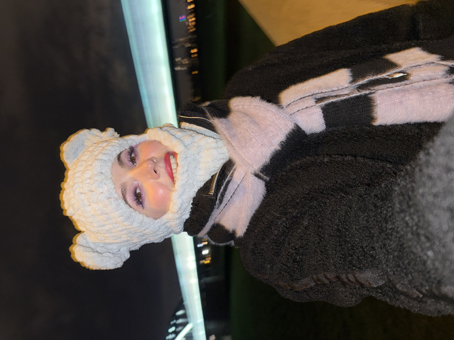 A woman wearing a white knit balaclava with bear ears smiles, looking towards the camera. She wears bright pink eyeshadow and lipstick. She is wearing a thick, dark textured coat with light purple and black faux fur accents. Behind her, there is a bright light source and a dark background.
