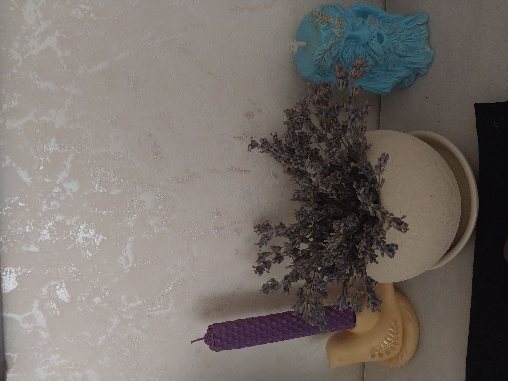 A still life arrangement is displayed on a light-colored surface against a textured off-white wall. From left to right, a yellow ceramic bird-shaped candle holder contains a tall, purple, honeycomb-patterned candle. To its right, a bouquet of dried lavender is arranged in a textured, cream-colored round pot with a saucer. To the right of the lavender, a blue, sculpted candle depicts a face with a beard and a leafy headdress. The overall scene is static, with no people or actions observed. The environment appears to be indoors, likely on a shelf or table.