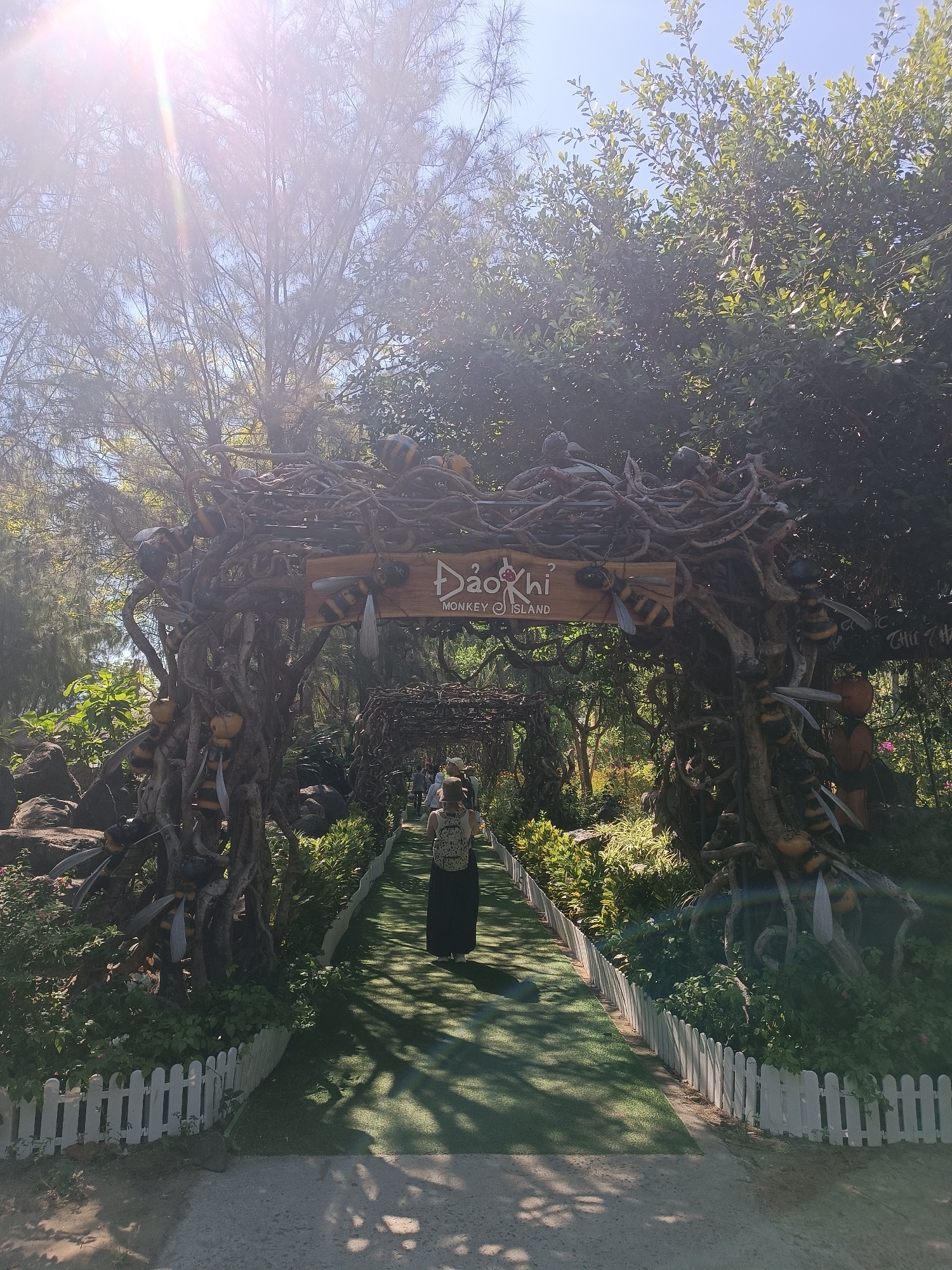 This media file depicts an outdoor scene with a pathway leading through a decorative archway adorned with large, stylized bee figures and intertwined branches. The archway is made of natural materials, appearing to be crafted from twisted roots and branches. The path itself is a vibrant green, possibly artificial turf, bordered by a low white picket fence.
A person, seen from behind, is walking along the path away from the viewer, wearing a backpack and a long dark skirt or dress. Other individuals are visible further down the path, also walking. The setting is lush and green, with abundant trees and foliage surrounding the pathway and archway, suggesting a park or natural attraction. The time of day appears to be morning or afternoon, with bright sunlight streaming through the trees, casting dappled shadows on the path. The sky is clear and blue.
A sign is visible on the archway with text that reads "ĐẢO KHỈ MONKEY ISLAND." This indicates the location as Monkey Island. The overall atmosphere seems serene and inviting, drawing visitors into a themed environment. The large bee decorations add a whimsical and playful element to the natural surroundings. The light flare in the upper left corner suggests a sunny day.