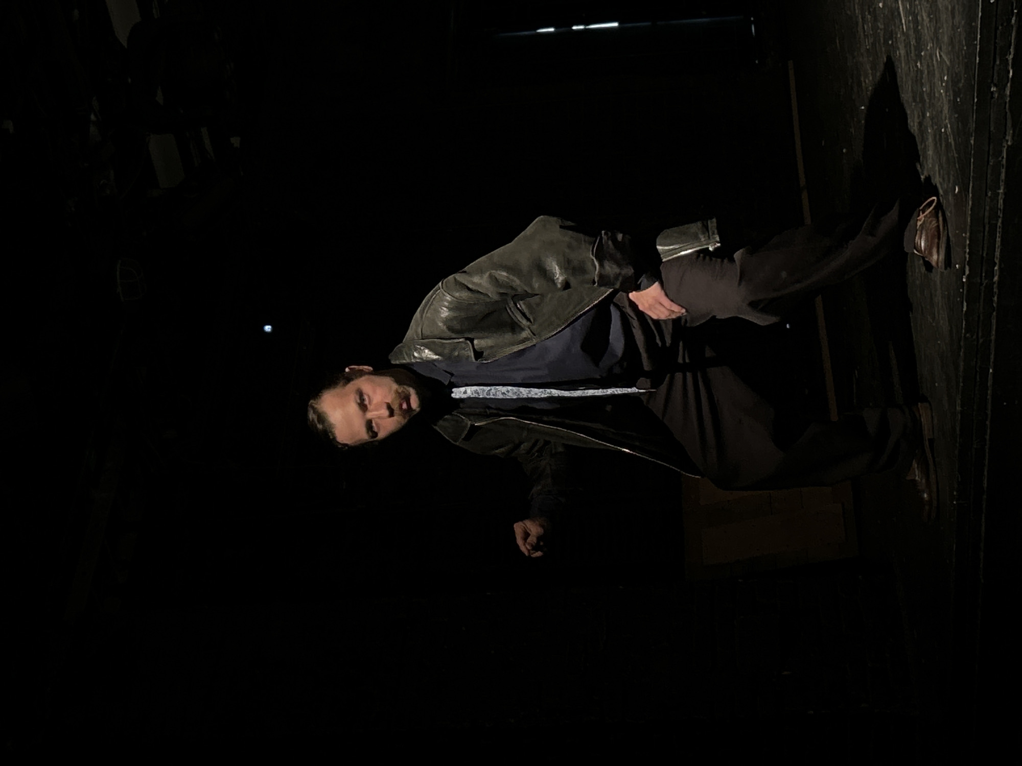 A man is standing in a dark room. He is wearing a leather jacket, black pants, and brown shoes. He is looking at the camera. The room appears to be a backstage area of a theatre or event space, given the dark curtains along the right side of the frame. The location is unknown, as the city is not specified.
