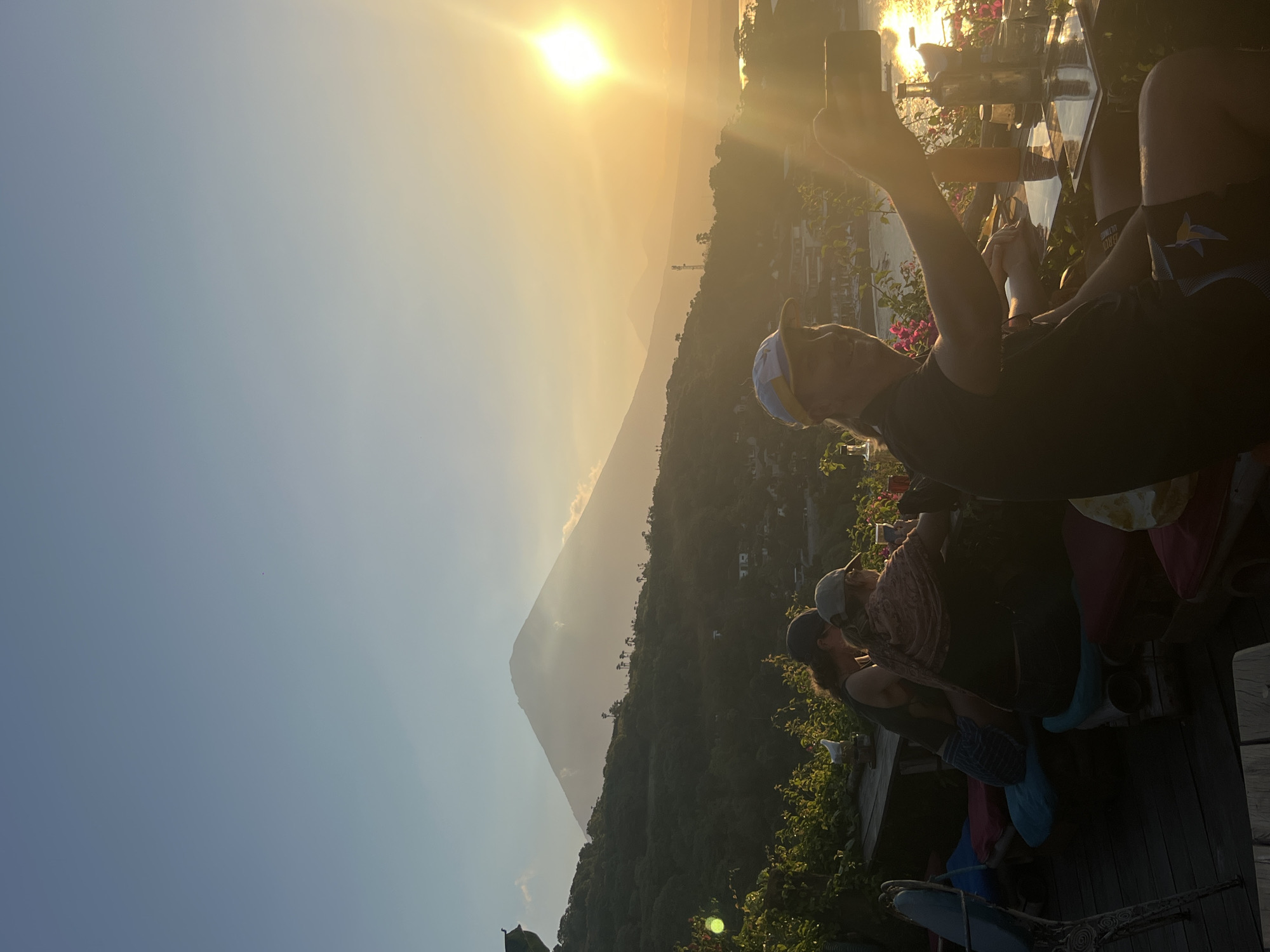 The image depicts a group of people gathered on a wooden deck, enjoying a sunset. The sun, a bright orb in a hazy sky, casts a warm golden glow over the scene. In the distance, a majestic mountain, possibly Mount Abang, rises from a verdant, tree-covered landscape. Buildings dot the hillside, suggesting a residential or resort area.
The foreground is dominated by the figures of several individuals, their faces partially obscured by the low angle of the sun and the orientation of the camera. One person in the center is holding up a smartphone, seemingly capturing the breathtaking view or taking a selfie. Another individual is wearing a baseball cap and a patterned scarf. People are dressed casually, with some wearing shorts. The overall mood appears relaxed and convivial, with the natural beauty of the sunset serving as the focal point. The wooden deck, adorned with plants, creates an inviting outdoor space. The time of day is clearly sunset, as indicated by the warm, low-angled sunlight. The weather appears clear with a slight haze, common in tropical settings. The presence of the mountain and the lush vegetation strongly suggest a tropical location, consistent with Abang, Indonesia. No visible text is present in the image.