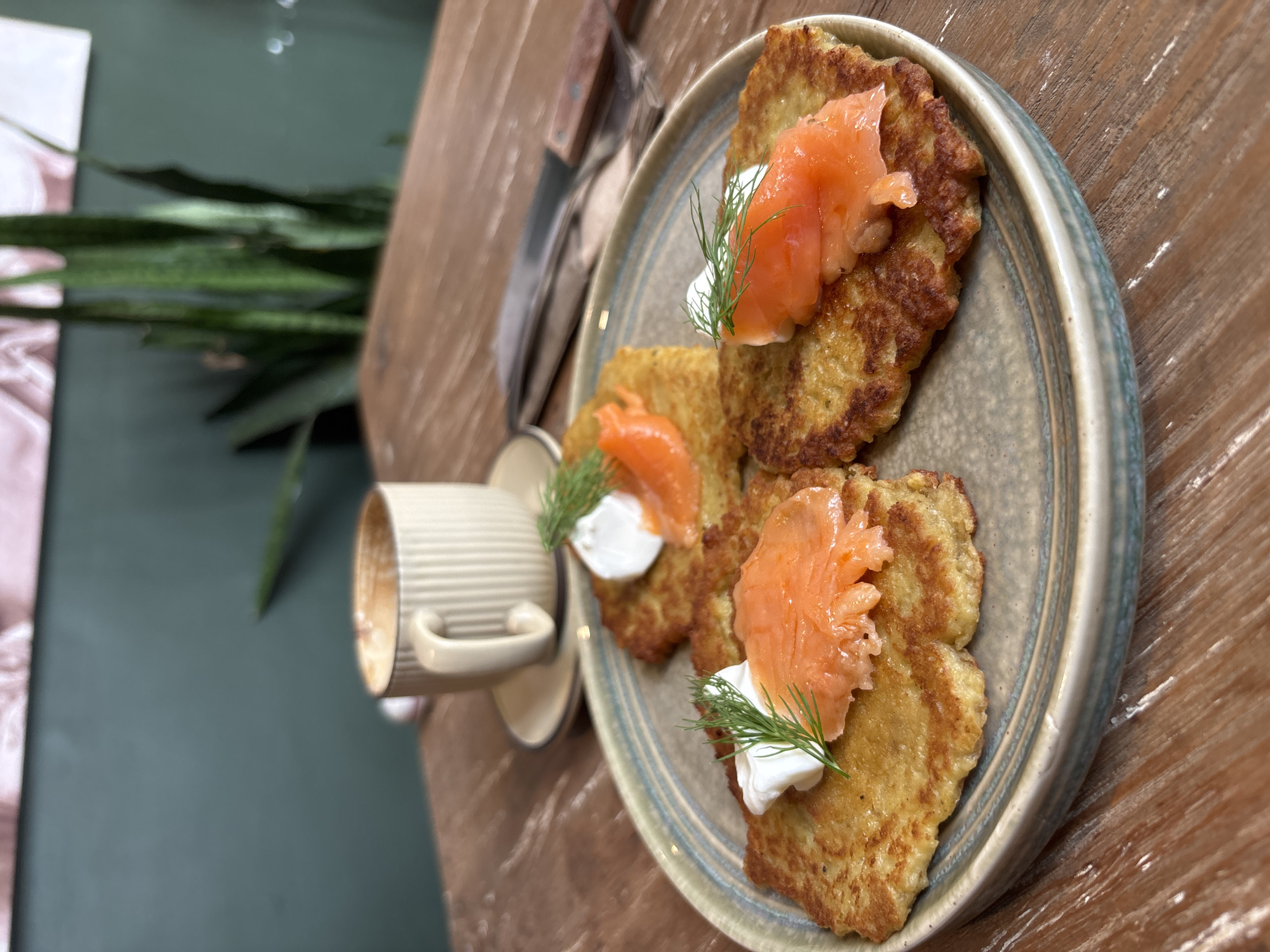 The scene depicts a plate of potato pancakes topped with smoked salmon, sour cream, and dill, alongside a cup of coffee, on a wooden table. The location is Kuta Selatan, Indonesia.