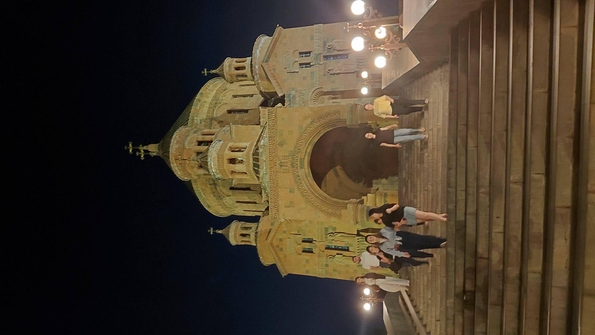 This media file shows a group of people gathered on a set of stone steps at night, with a large, intricately designed building in the background. The building appears to be a church or cathedral, with multiple domes and ornate carvings. It is illuminated by exterior lights, casting a warm glow. The sky is dark, indicating it is nighttime. The people are positioned at various levels on the steps, some standing, some sitting. They are dressed casually in a mix of light and dark clothing. One person in the upper portion of the steps is wearing a yellow shirt and dark pants. Another person below them is wearing a black top and jeans. A group of younger individuals are further down, some appearing to be interacting with each other, suggesting a social gathering or a sightseeing visit. The overall mood seems relaxed and convivial. The steps are wide and made of large stone blocks. Decorative lights, resembling old-fashioned street lamps, are visible on poles to the left of the steps, also illuminated. The architecture of the building and the style of the lights suggest a location in Eastern Europe or the Middle East, consistent with the provided location context of Abovyan, Armenia. There is no visible text in the image.