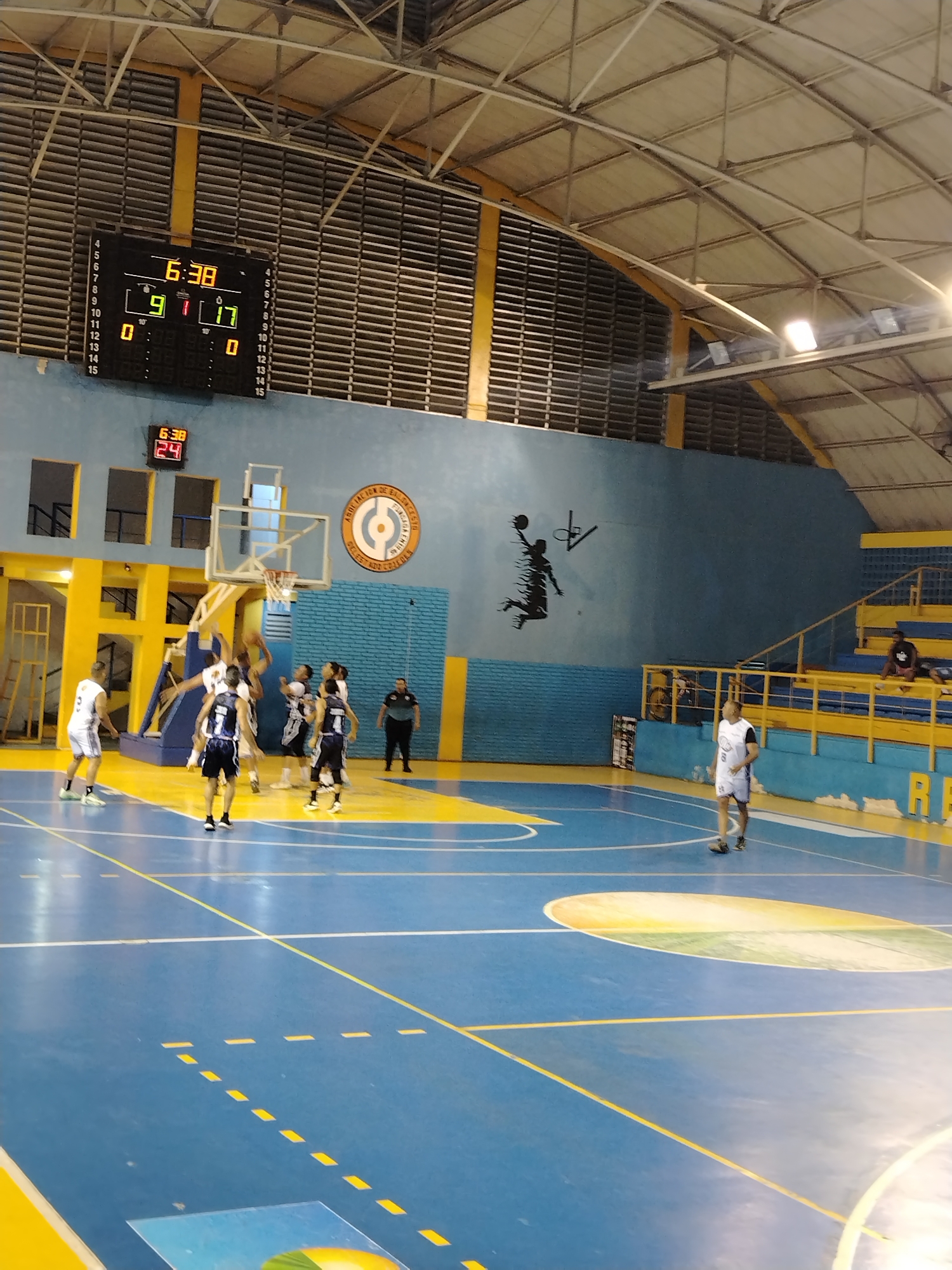 The image depicts a basketball game in progress inside a gymnasium. The scoreboard shows a time of 6:38 and a score of 9 to 17. Several players in white and blue uniforms are actively engaged in a play near the basketball hoop. An official stands on the sidelines. Spectators are seated in the bleachers. The court is blue with yellow accents. A silhouette of a basketball player dunking is visible on the wall. A logo is featured on the blue wall. The gymnasium has a metal roof and louvered windows. Based on information available online, it is highly probable that this gymnasium is located in Tinaquillo, Cojedes, Venezuela.