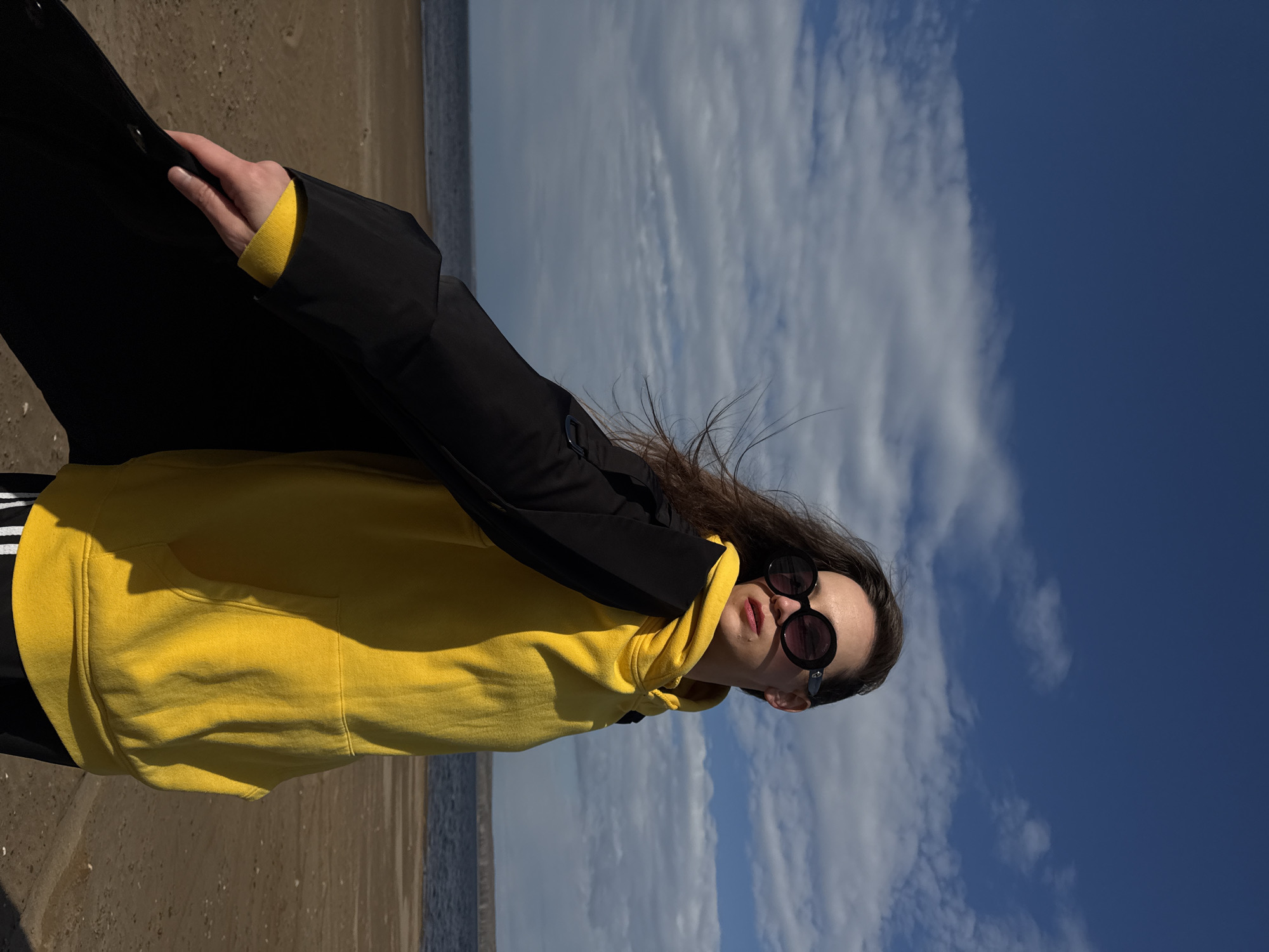 A young woman stands on the sandy beach of the Votkinsk Reservoir in Chaikovsky, Russia. She wears a yellow hoodie with three white stripes on the sleeve, a black coat, and round sunglasses. The background is a mix of a cloudy sky and the reservoir's shoreline.