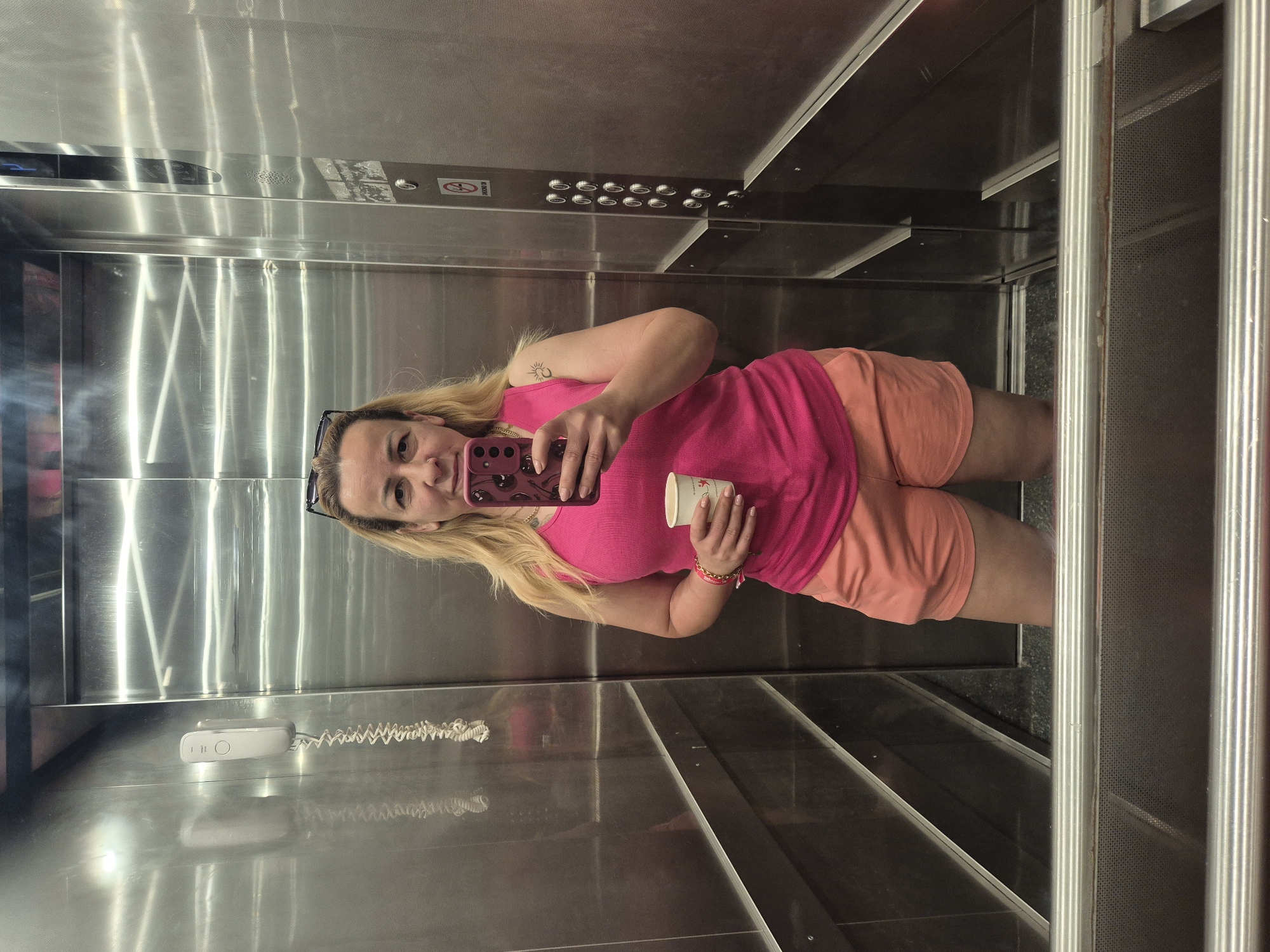 In an elevator, a woman with blonde hair tied up in a ponytail stands facing a mirror, taking a selfie. She is wearing a bright pink tank top and coral-colored shorts. In her right hand, she holds a white paper cup, and in her left hand, she holds a pink smartphone with a cherry-patterned case. She is also wearing sunglasses on top of her head and several bracelets on her left wrist. The elevator walls are made of stainless steel and reflect the lighting. On the left wall, an intercom system is mounted, and on the right wall, an elevator control panel with buttons is visible. The floor of the elevator is dark and appears to be made of tile or stone. A metal handrail runs along the bottom of the frame.
