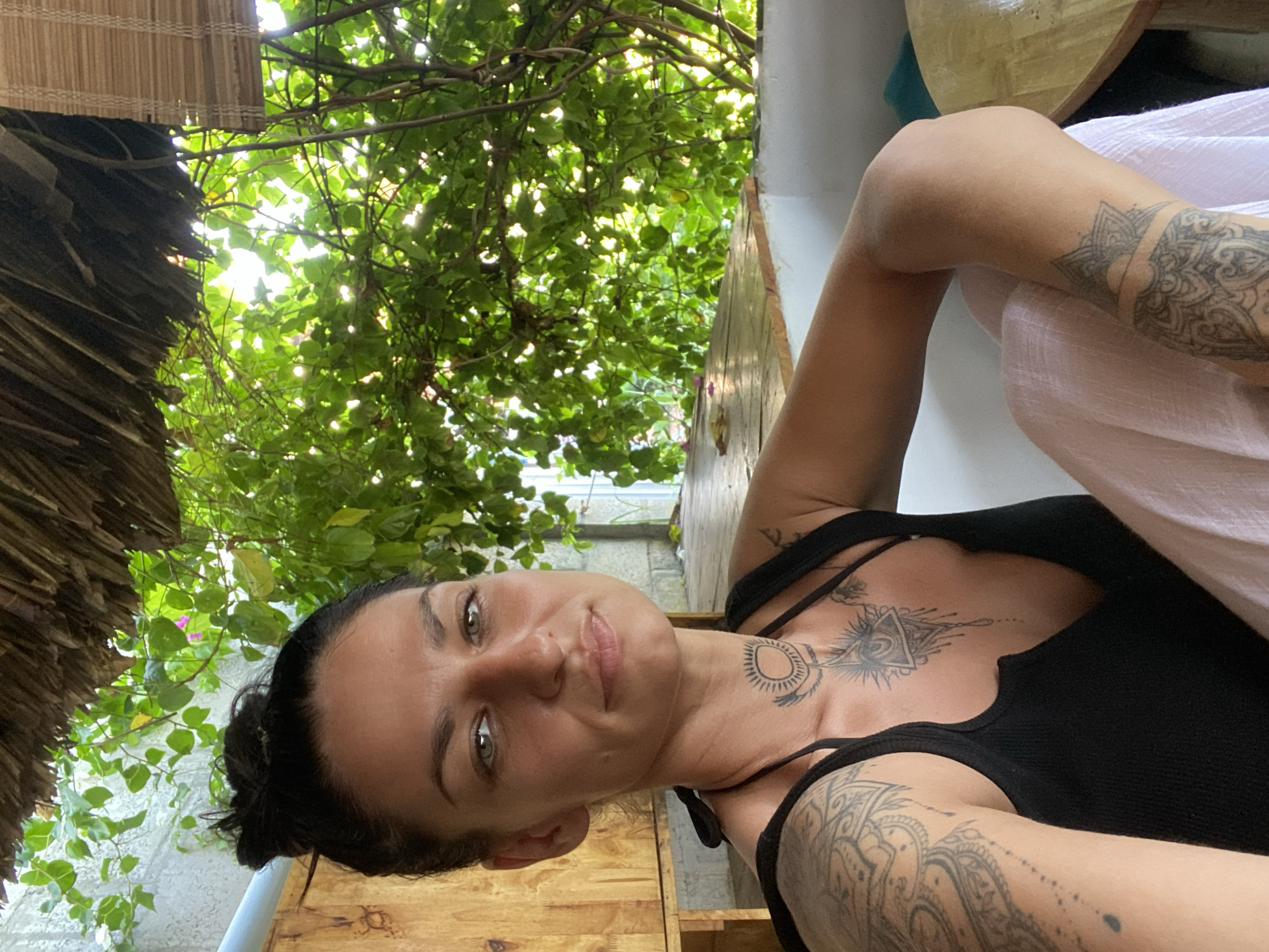 This image depicts a woman seated in a semi-outdoor or open-air structure located in Phu Thuy Ward, Vietnam. The woman, positioned in the lower-left to central portion of the frame, has dark hair styled in a bun, green eyes, and a composed expression while looking directly at the camera. She is wearing a black tank top with a deep V-neck. Multiple tattoos are visible: an extensive mandala-style pattern covers her left upper arm and shoulder; a circular design resembling a sun with rays and two dots is situated on her throat; an Eye of Providence within a triangle, detailed with geometric and hanging elements, is present on her sternum; and an intricate geometric pattern adorns her right forearm. Her right arm is bent, resting over her lap, which is covered by light-colored fabric.
The background elements suggest a natural and rustic environment. To the viewer's left, a light-colored wall is partially covered by green climbing foliage featuring small white and pink flowers. Below this, a light-colored wooden structure is visible. A thatched roof extends across the top of the frame. The right and upper-right background is dominated by dense, vibrant green foliage, through which bright sunlight filters, creating luminous spots. A woven bamboo or wooden blind is partially visible on the far right. A light-colored wooden surface, consistent with a table or ledge, is present in the mid-ground on both sides of the woman, and a white ledge is visible in the lower right. The scene is illuminated by strong natural daylight, with prominent backlighting emanating from the dense foliage.