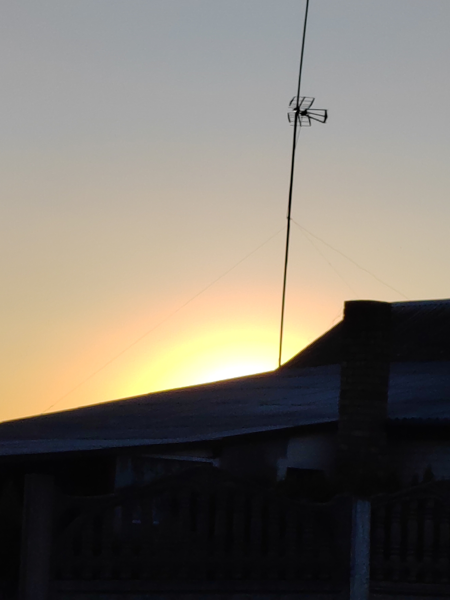 This image depicts a silhouetted rooftop and fence against a pale orange and yellow sky, suggesting either sunrise or sunset. A tall, thin antenna with a directional element at its top stands prominently against the sky. The sky itself is a gradient, transitioning from a lighter hue at the top to brighter, warmer tones near the horizon. The rooftop is dark and appears to be corrugated metal. A brick chimney or structure rises from the roof. In the foreground, a dark, silhouetted fence with vertical posts is visible. The overall scene evokes a quiet, possibly rural setting. The light suggests the early morning or late evening. The provided location context of Воўчынскі сельскі Савет, Belarus, places the scene in a specific geographic region. There is no visible text in the image.