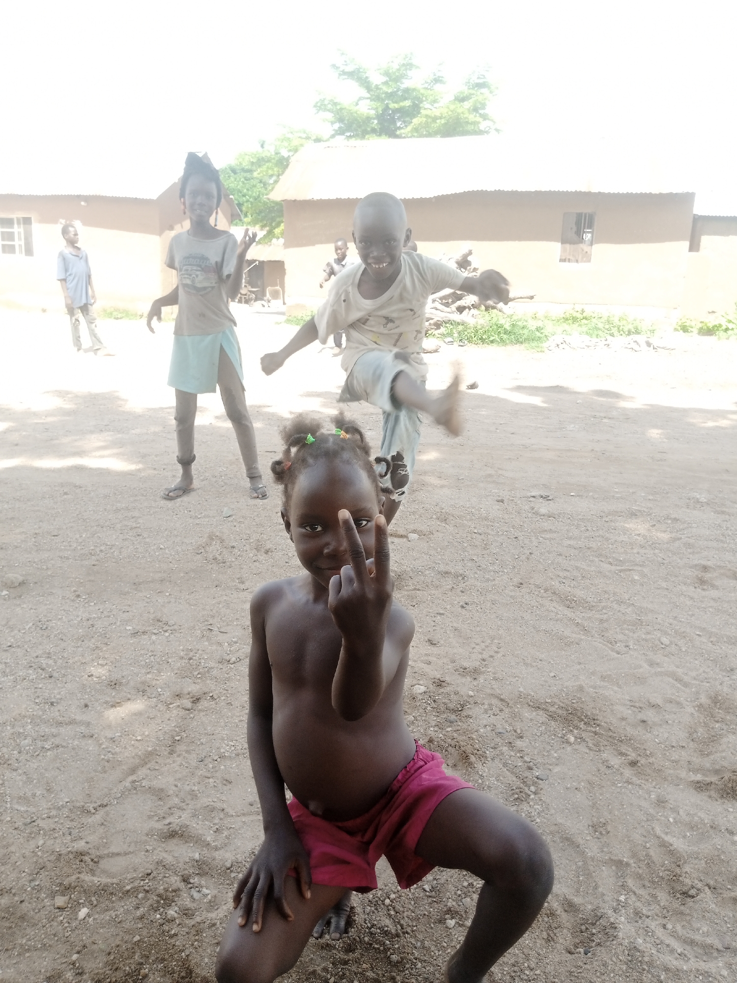 A group of four children are in an outdoor, dusty area in front of two mud-brick buildings with thatched roofs. The child closest to the camera, a young girl with dark skin and braided hair adorned with colored beads, is in the foreground. She is shirtless and wearing red shorts, and is sitting with her left knee bent and her right leg extended. Her left hand rests on her bent knee, and her right hand is raised, with her index and middle fingers extended and pointed towards the camera. Behind her, to the left, is another child, a girl with dark skin and dark hair, wearing a light blue skirt and a grey t-shirt with a graphic print. She is standing with her arms outstretched and her head tilted back as if playing. To the right of her, and further back, is a boy with dark skin, shirtless and wearing light-colored shorts. He appears to be mid-jump or kicking, with his right leg extended forward and his left leg bent. Another child, a boy with dark skin, is visible in the far left background, wearing a blue t-shirt and pants. The ground is sandy and sparsely covered with small stones and some green vegetation. The sky is bright and blown out, suggesting it is a sunny day.