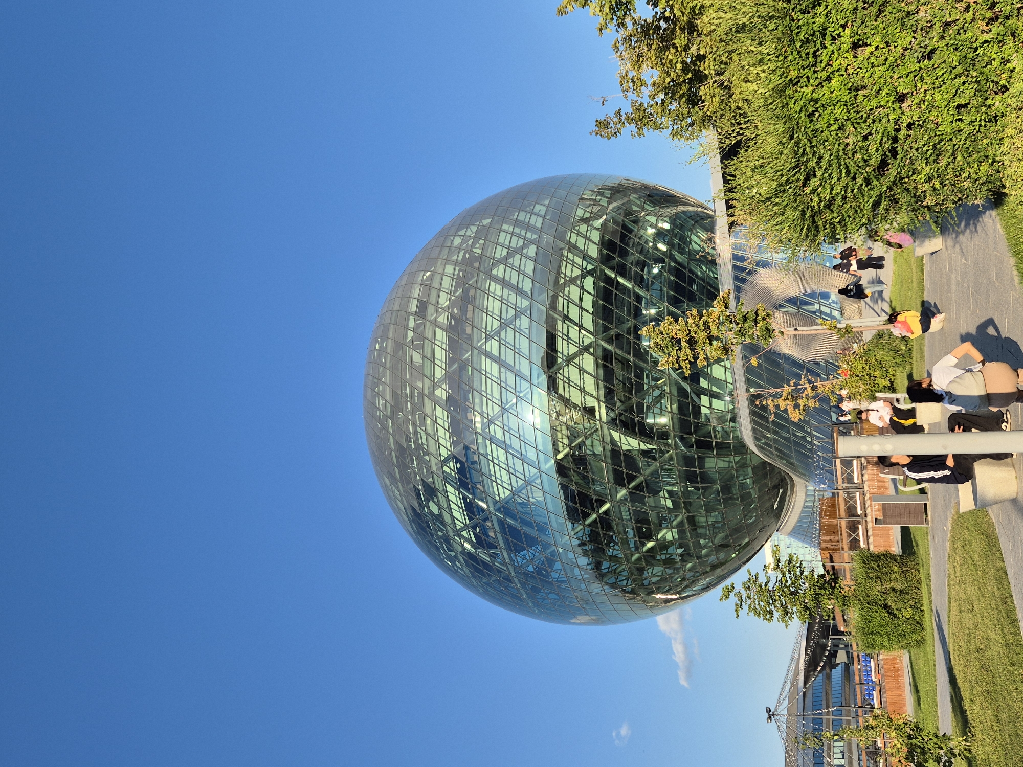 The image shows a large, spherical glass building, reflecting the bright blue sky. The building has a geodesic dome structure with triangular glass panels. In front of the building, there is a park with green grass, trees, and bushes. Several people are sitting on benches in the park, some interacting with each other. The scene appears to be an outdoor public space in Astana, Kazakhstan. The building is likely the Nur Alem Pavilion, also known as the Future Energy Museum, a prominent landmark in Astana.