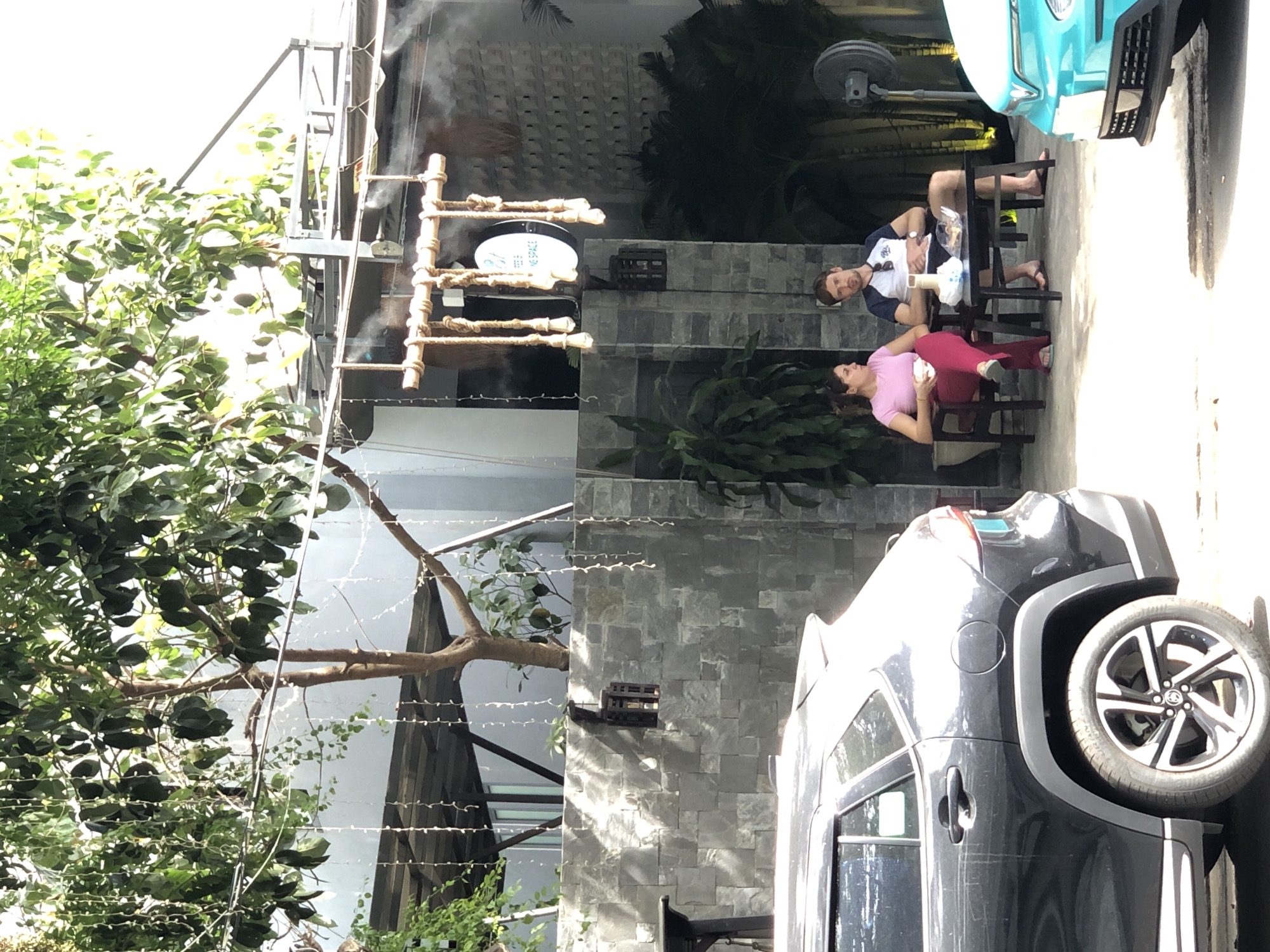A couple sits at a table outside a cafe in Da Nang, Vietnam. The woman, wearing a pink t-shirt and red pants, holds a white cup. The man, in a blue and white shirt and black shorts, sits with his arms crossed. A black SUV is parked to the left, and a bright teal car is parked to the right. The cafe has a grey stone wall with a hanging lantern and decorative string lights. In the background, a tree with lush green leaves shades the area, and mist is being sprayed from pipes, likely for cooling.