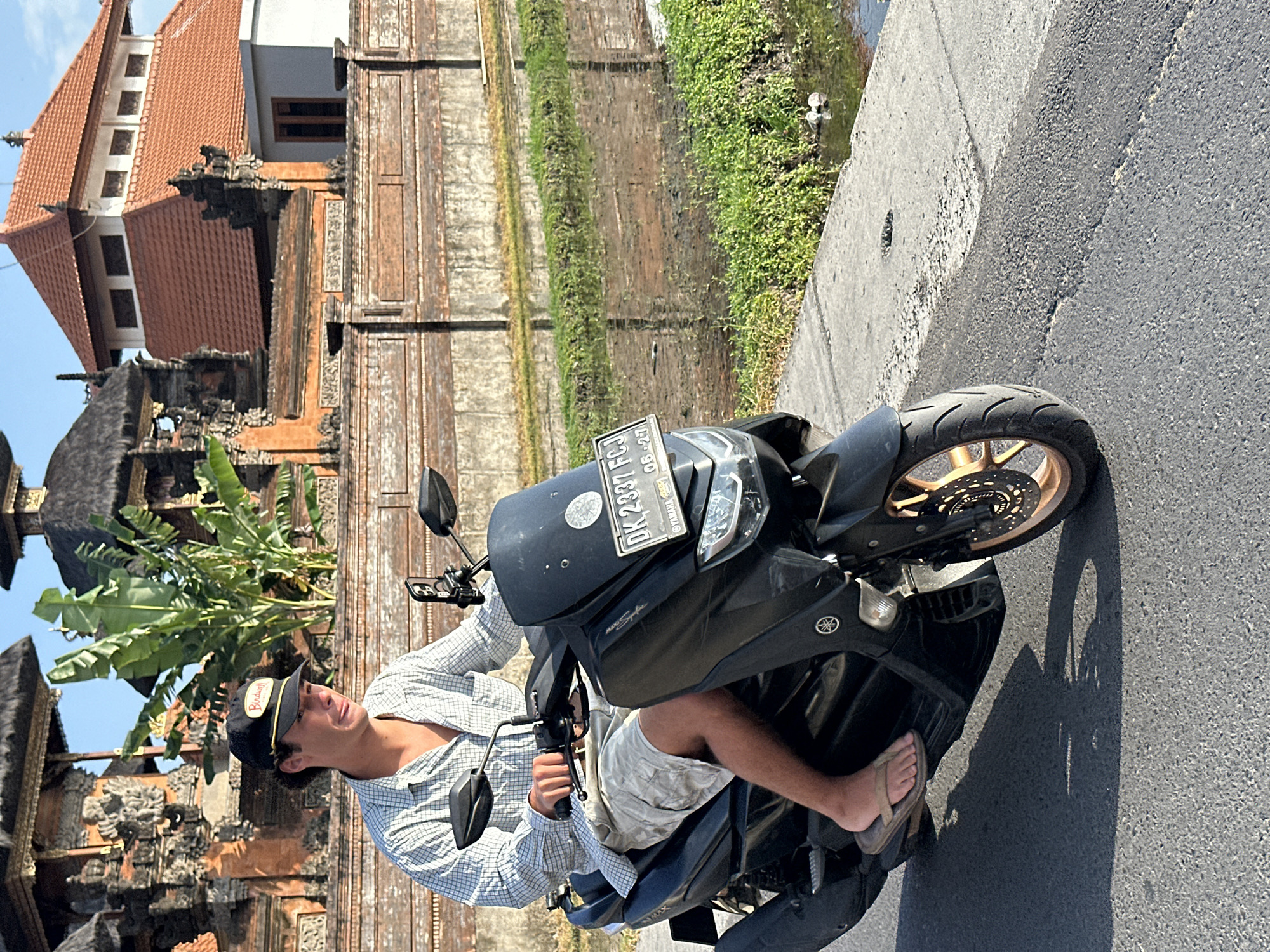 A young Caucasian male wearing a plaid shirt, grey shorts, and a black baseball cap with a "Birdwell" logo is riding a black scooter on a paved road. He is looking down and to his left with his mouth slightly open. The scooter has a license plate that reads "DK 2337 FCJ 05-27". In the background, there are traditional Balinese buildings with ornate stone carvings and terracotta roofs, and a rice paddy with lush green vegetation. The sun is shining brightly, casting shadows on the road. The overall scene depicts a cultural setting in North Kuta, Indonesia, with a focus on the individual's journey through this landscape.