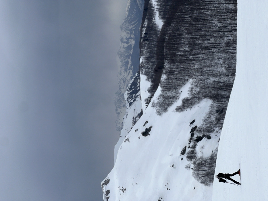 An elevated winter landscape view at Katun Bačko brdo, Montenegro, under a heavily overcast, dark grey sky. The scene is dominated by snow-covered mountains and slopes. In the lower-left quadrant, a single individual is observed on a snow-covered slope, positioned slightly right of the frame's center. The person is dressed in dark winter clothing, including a black jacket and pants, with a light-colored helmet, and is equipped with skis and ski poles. They appear to be in a downhill skiing stance. Behind the skier, a vast expanse of snow-covered terrain rises, featuring scattered rocky outcrops and clusters of bare deciduous trees. To the right and extending into the midground, a dense forest of leafless trees covers a steep hillside, contrasting sharply with the white snow. In the background, a range of snow-capped mountains with rugged peaks is visible, partially obscured by atmospheric haze. The overall lighting is diffused, indicating an overcast condition.
