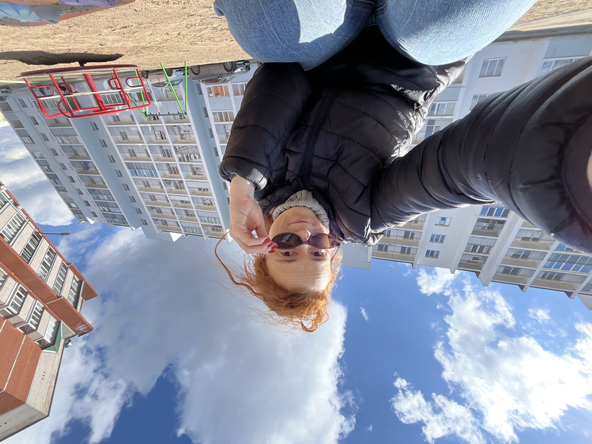 The original image is presented rotated 90 degrees counter-clockwise. Described here in an upright orientation, the photograph is a low-angle outdoor selfie featuring a woman looking up into the camera. She has red or auburn hair and is dressed in a black puffer jacket, a light grey knit beanie, and blue jeans. With her right hand, she is pulling down her dark sunglasses, revealing her eyes, while her left hand is also partially visible. Her expression appears neutral or slightly inquisitive.
The background shows a bright blue sky with scattered white, fluffy clouds, indicating a sunny day. Below the sky, several multi-story residential buildings are prominent; a large, light-colored apartment building with numerous balconies extends across the right and center, while a building with orange-brown and grey sections stands on the left. In the mid-ground, a red metal merry-go-round and some green playground structures are discernible, with parked cars behind them. A patch of sandy or light brown ground is visible in the immediate foreground, surrounding the woman.
The scene depicts a typical urban or suburban setting, likely a playground or courtyard of an apartment complex in Chaikovsky, Russia. There is no visible text in the image.