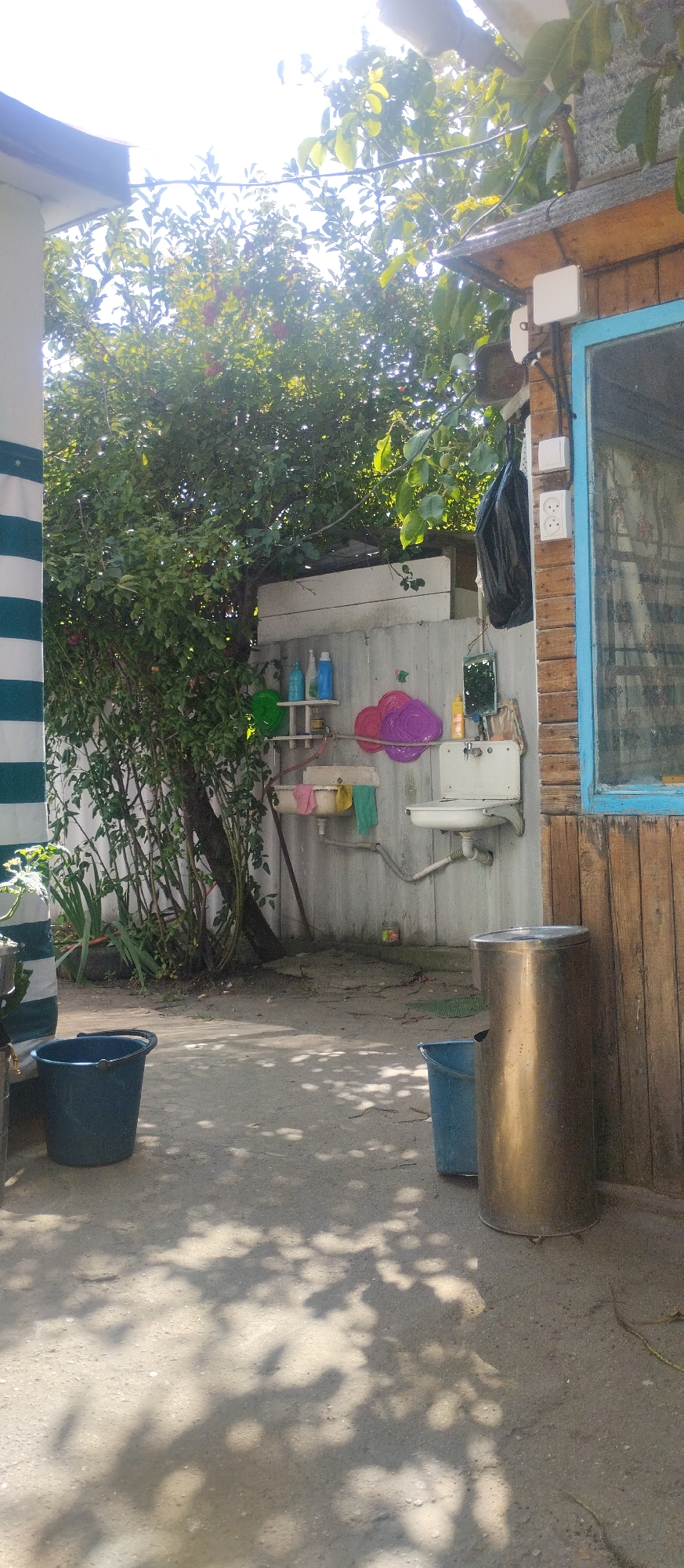 The image depicts an outdoor area in Cholpon-Ata, Kyrgyzstan, featuring a rustic sink setup against a corrugated metal wall. To the left, a large green bucket sits on the ground. Behind it, a lush green tree with red berries partially obscures the scene. The sink is white, mounted on the wall, with plumbing visible. Above the sink, shelves hold several bottles of cleaning products. Two colorful plastic plates, one purple and one pink, are hung on the wall. A small mirror is also present. A trash bag hangs from a hook above the sink area. To the right of the sink, a large, metallic trash can stands next to a blue bucket. A wooden structure with a blue-framed window is visible to the far right. The ground is a paved surface with patches of dirt, and dappled sunlight casts shadows from overhead foliage. The overall impression is one of a functional, outdoor utility area in a residential setting.