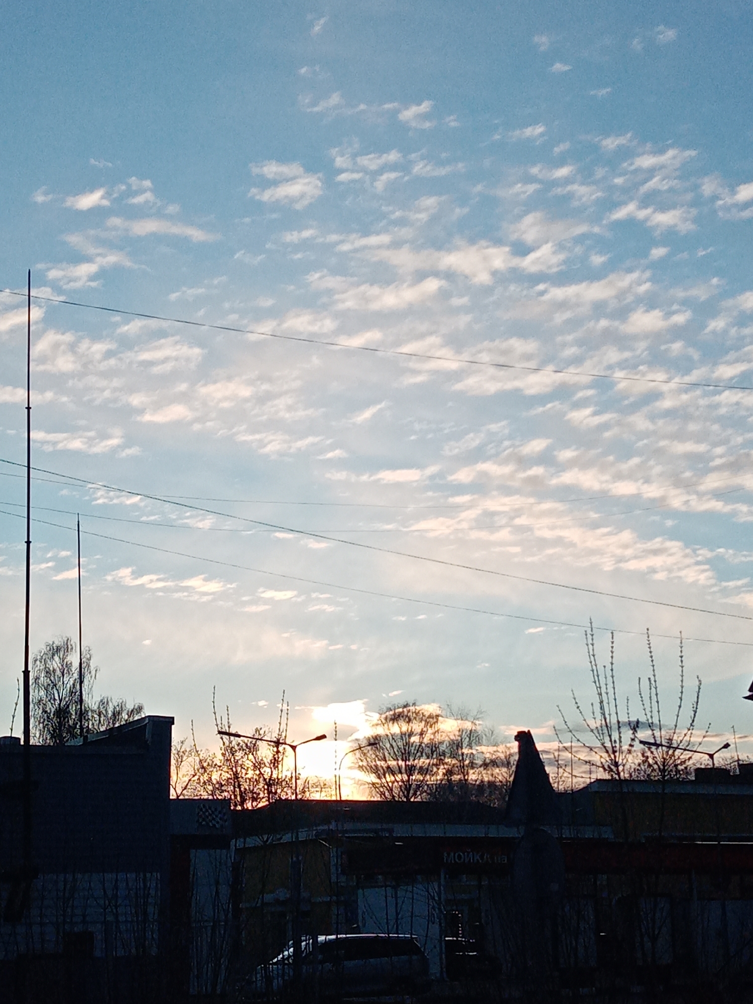 The image shows a view of Elektrostal, Russia at either sunset or sunrise. The sky is mostly blue with light white clouds scattered across it. Electrical wires run horizontally across the frame. In the foreground, silhouetted buildings, trees, and street lights obscure the lower part of the image. The word "МОЙКА" (car wash) is visible on a sign on one of the buildings. The sun is positioned low on the horizon, partially hidden behind the buildings and trees. Two tall antennas or poles are visible on the left side of the frame. A car is parked in front of the buildings.