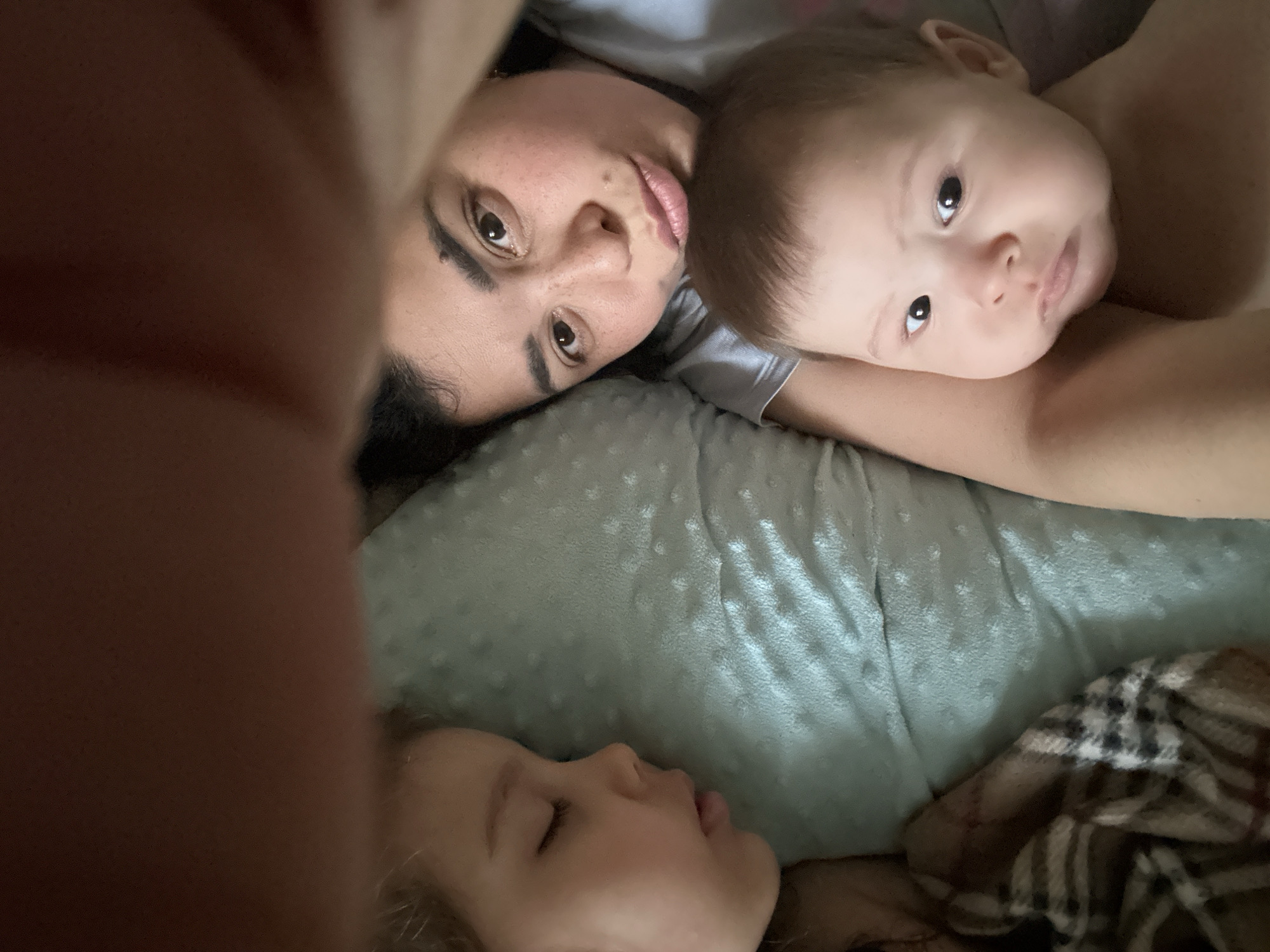 A selfie-style photograph features a woman and two children, presumably her children, lying together on a bed or similar soft surface. The woman is facing the camera with her head turned slightly, while the younger child on her right is also looking directly at the camera. A toddler is lying on the bottom of the image with their face in profile. The background consists of a soft, light green textured blanket or sheet and a plaid blanket. Part of the photographer's hand is visible in the lower left corner. The location is Unknown, Unknown.