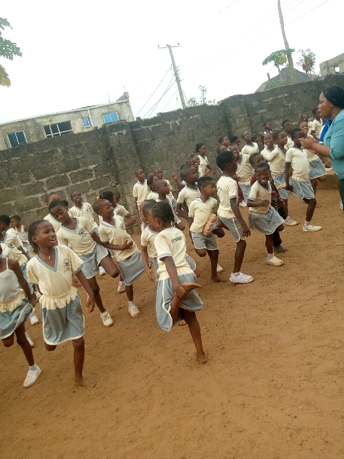 The image captures an outdoor scene in Ukwu-Abwa, Nigeria, featuring a group of approximately 30 to 40 primary school-aged children engaged in an activity, supervised by one adult female. The children are dressed in a uniform consisting of light-colored, short-sleeved tops with V-necks and light blue or grey skirts/shorts, some with a crest on the chest. Some children wear white athletic shoes, while others are barefoot. The adult female, positioned on the far right, wears a light blue short-sleeved top and faces the children, gesturing with her hands.
The children display various poses indicative of physical activity; several have one leg bent at the knee with the foot lifted, some holding their ankle, suggesting stretching or a dance movement. Many children are smiling and looking towards the adult. The ground is composed of reddish-brown dirt. In the background, a high wall constructed from grey concrete blocks runs horizontally across the frame. Beyond this wall, multiple buildings are visible, including a multi-story structure with blue-tinted windows. Overhead, utility poles with numerous power lines are visible against an overcast sky. Sparse tree foliage is present in the upper left corner.